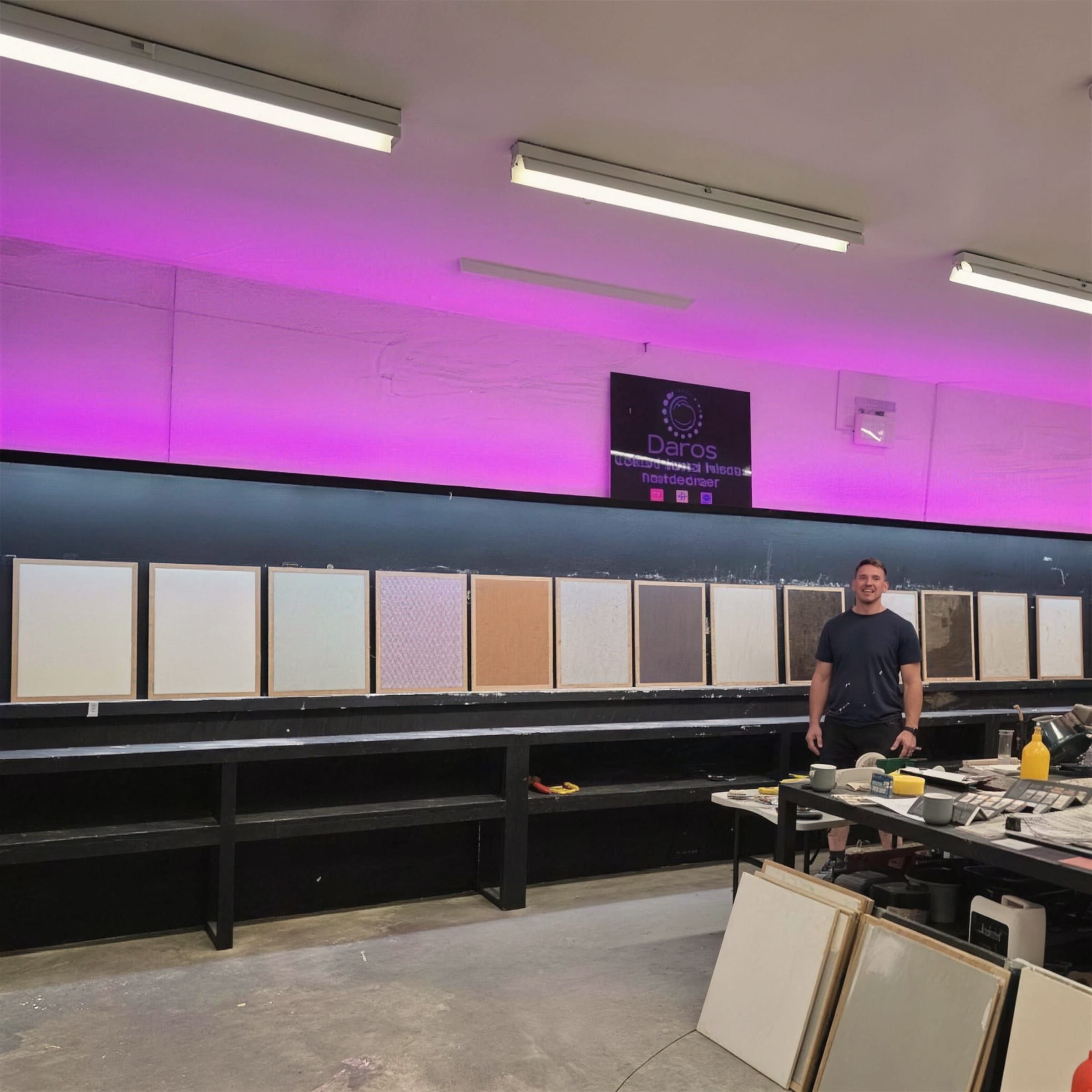 A long row of fresh sample boards lines the blackboard wall of the workshop under magenta light, Darren standing mid-frame by the workbench.