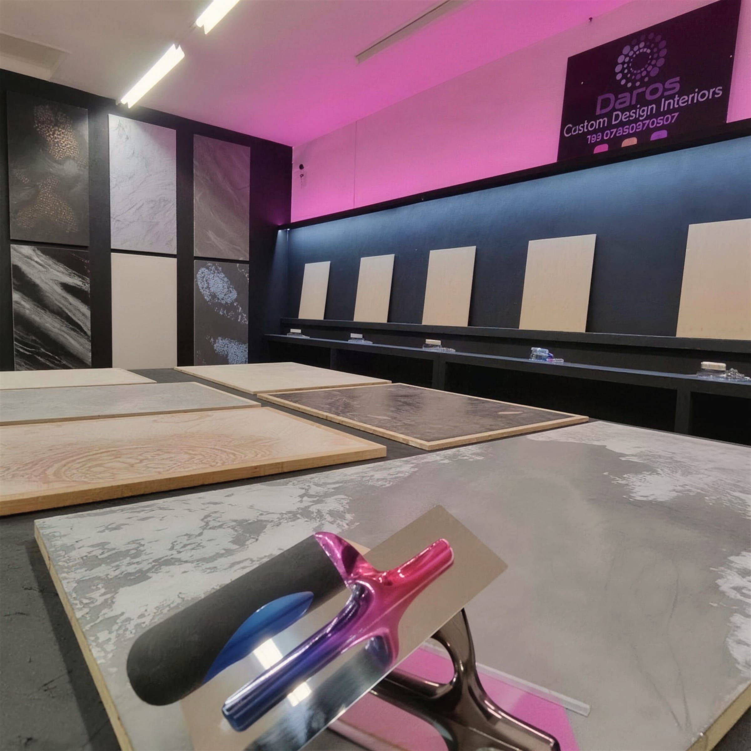 A chrome trowel resting on a bench of textured sample boards in the showroom, magenta LED light washing the wall behind