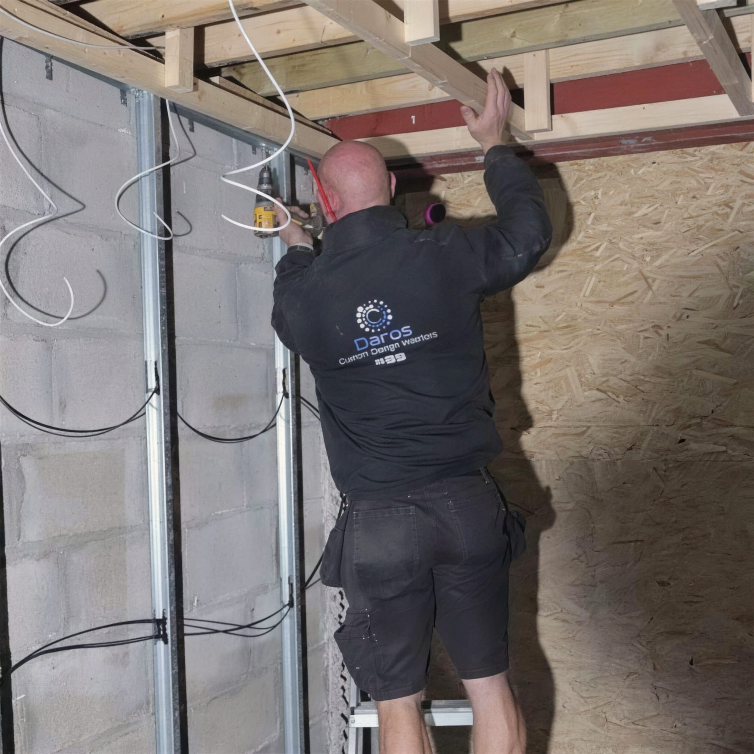 A Daros-jacketed worker on a ladder fixes into timber joists above metal stud framing, cables coiled against the blockwork wall.