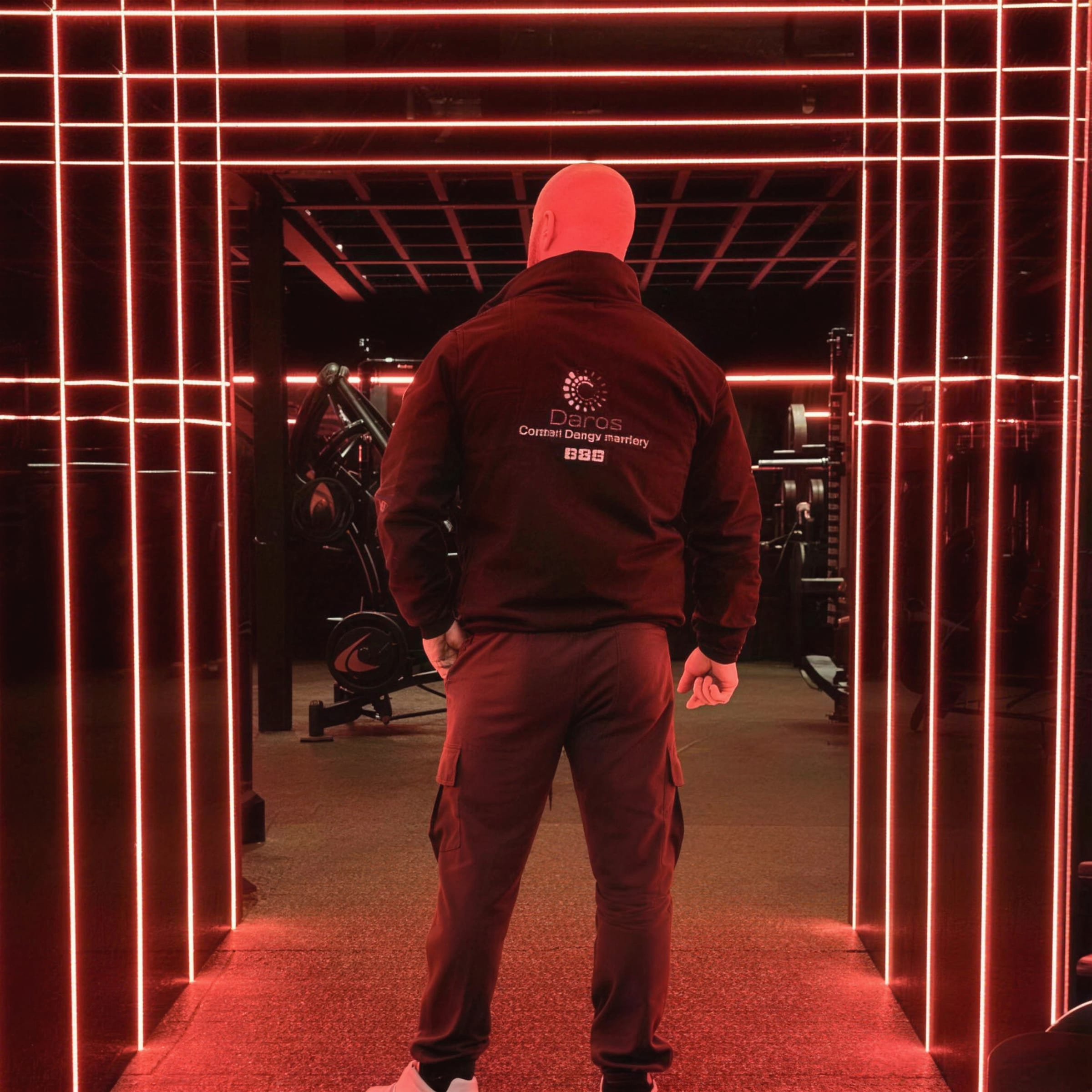 Daros in a DAROS hoodie standing inside the red linear-LED tunnel of a gym fit-out, the light grid receding behind him.