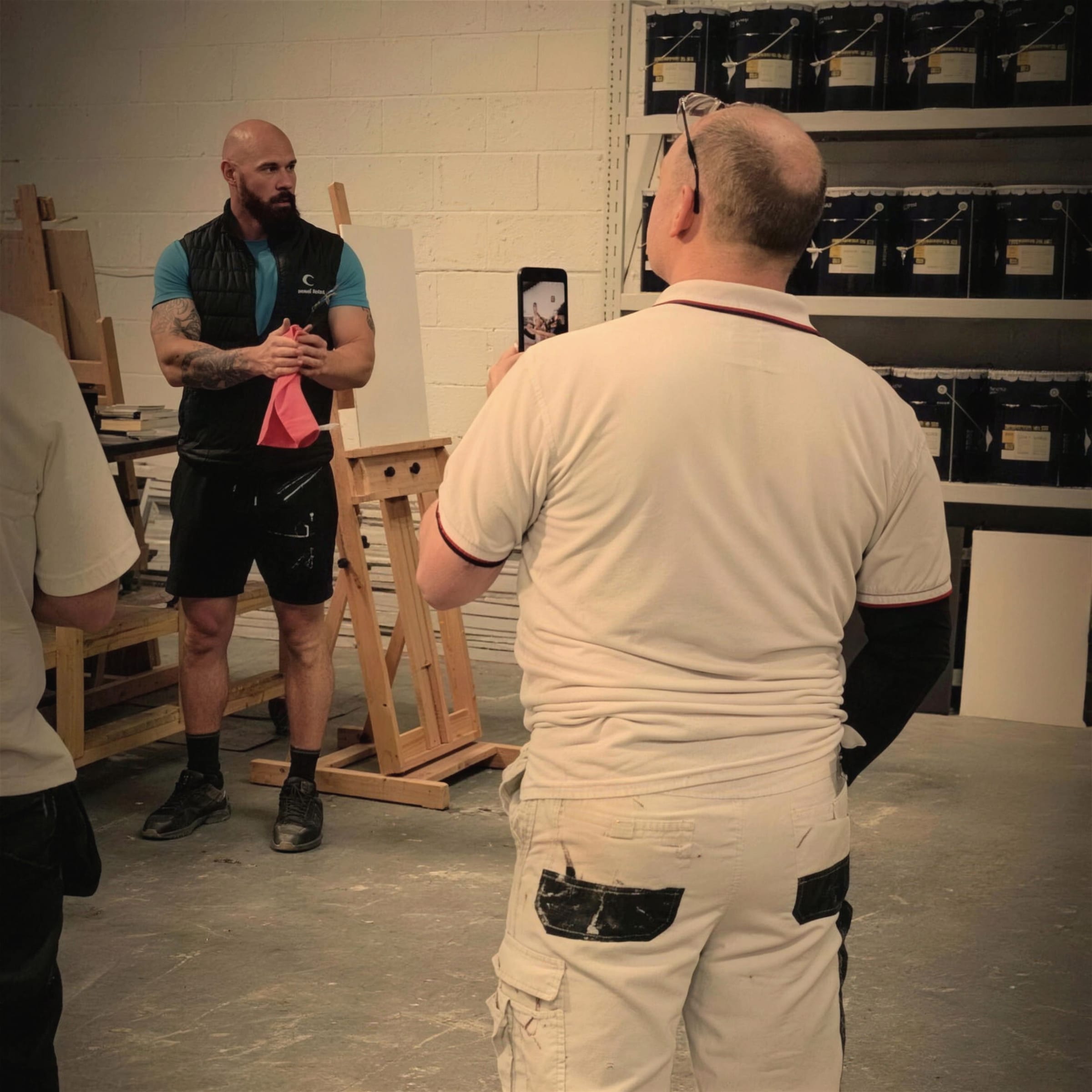Darren addresses a group of students in whites in the school workshop, racks of tins behind him, one student filming on a phone.
