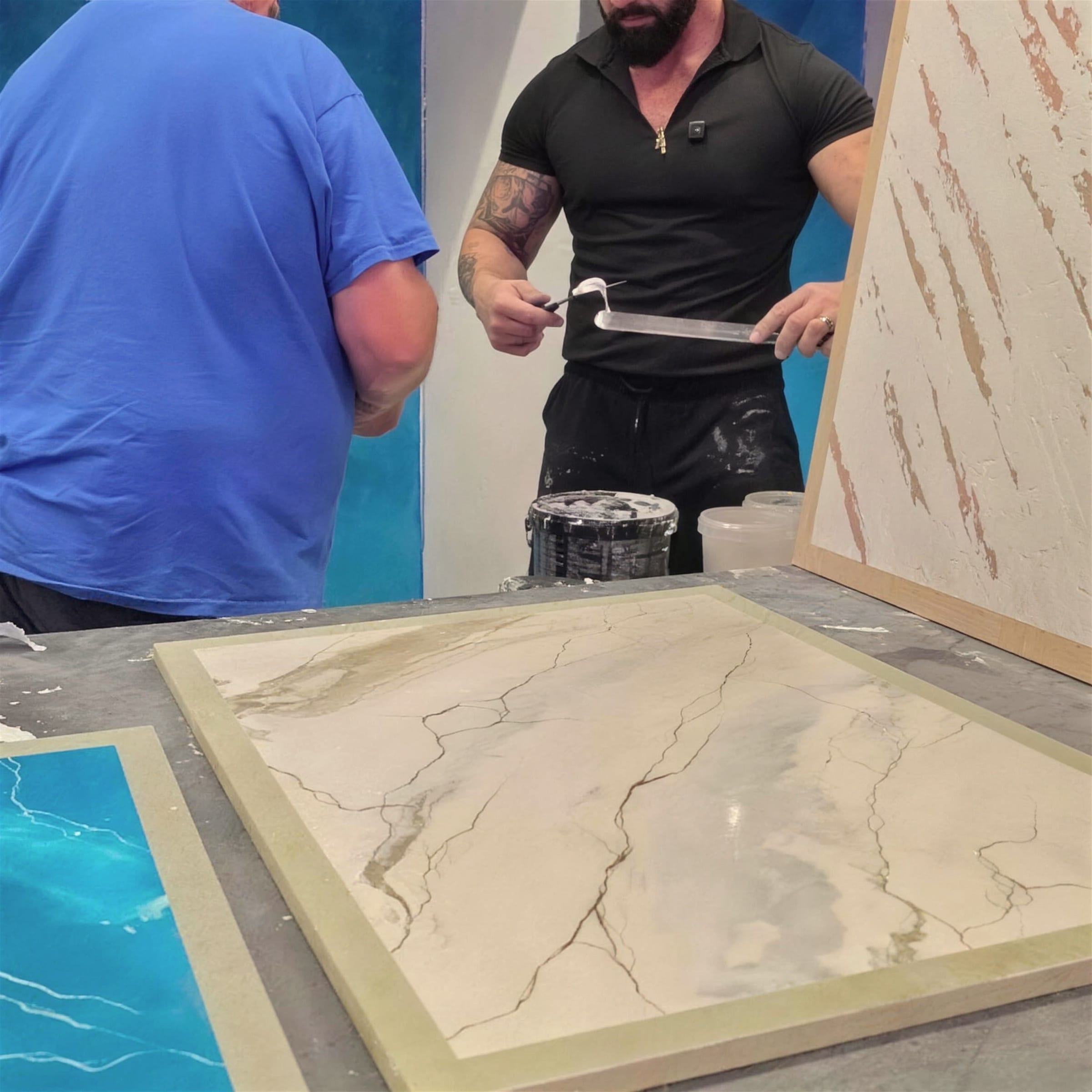 Bench-level view of a cream marble-effect training panel, the instructor cleaning a long blade above it and teal sample boards beyond.