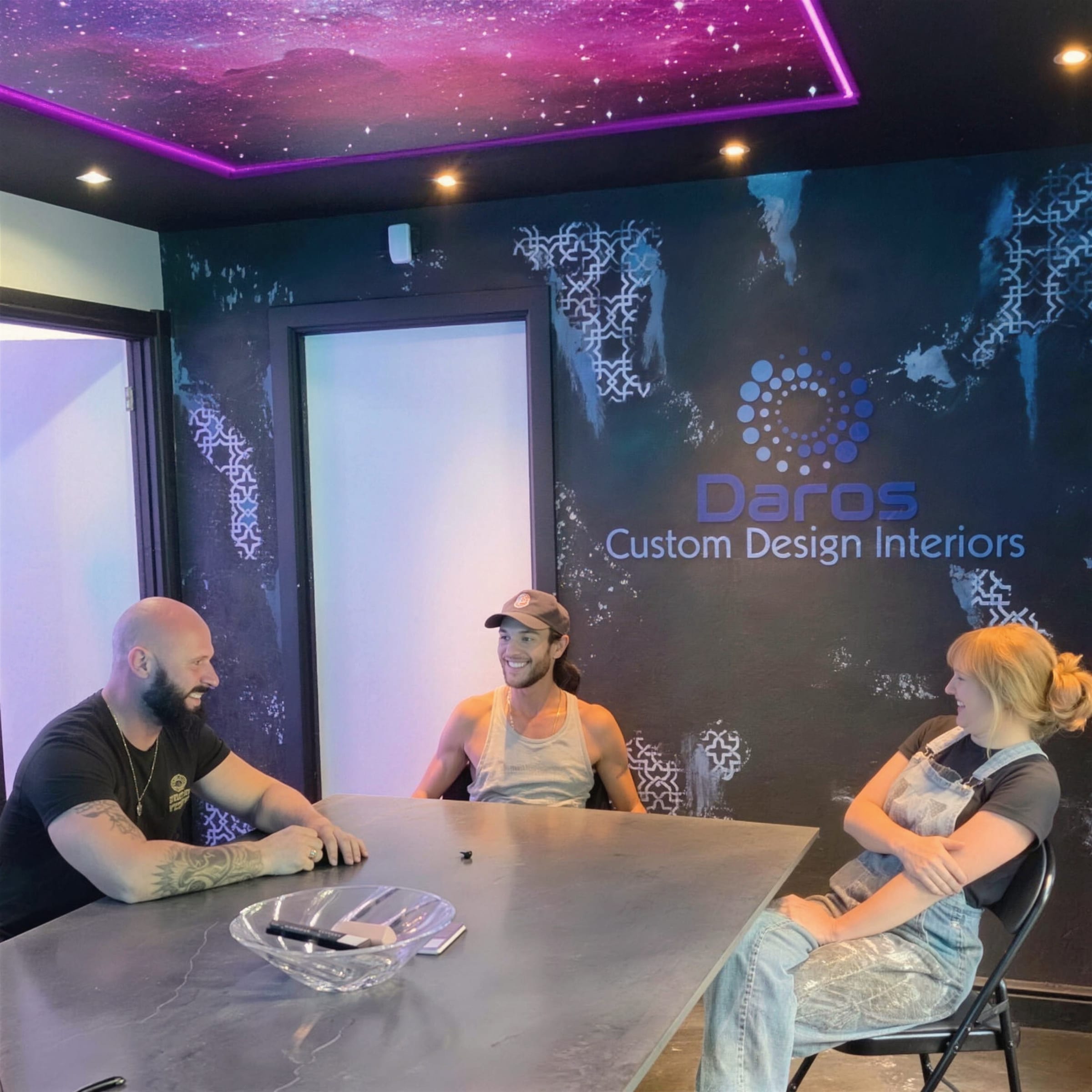 Darren and two trainees sit around a steel-topped table in the showroom, a galaxy-print ceiling edged in violet neon overhead and the Daros wall lockup behind.