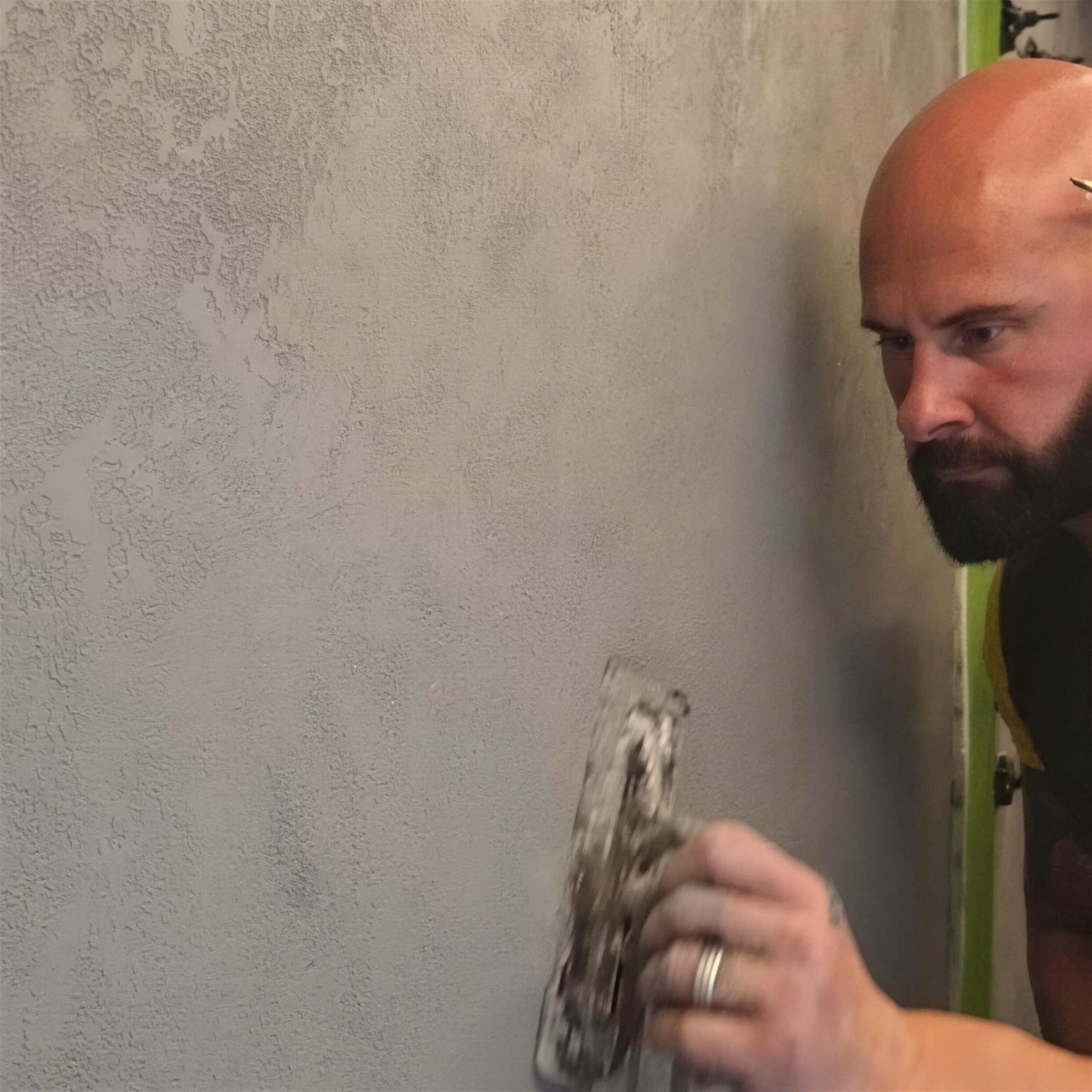 Darren studies the wall as he floats a loaded trowel over a stippled grey render coat, head cocked toward the surface.