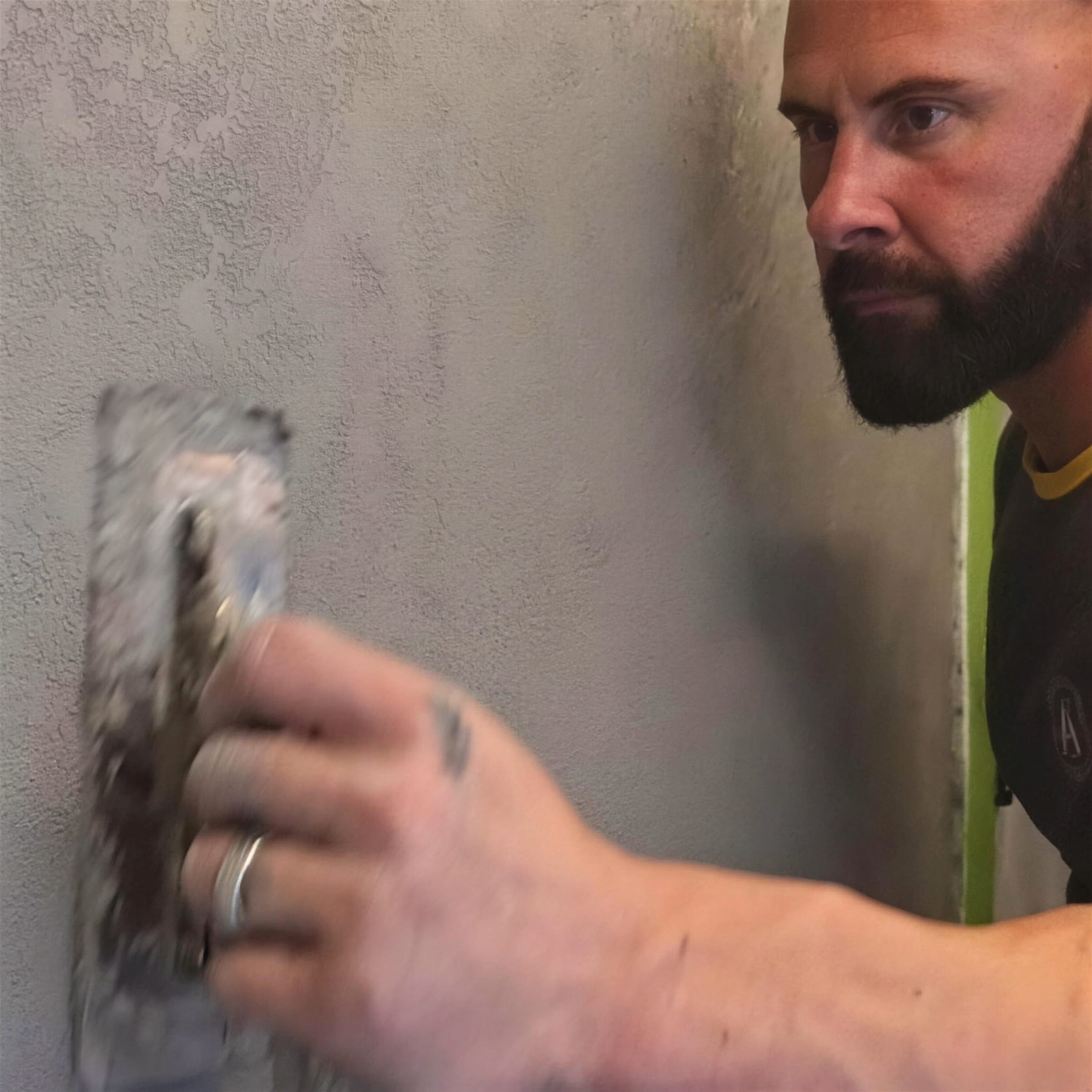 Close profile of Darren drawing a plaster-laden steel trowel across a freshly textured grey wall, eyes fixed on the surface.