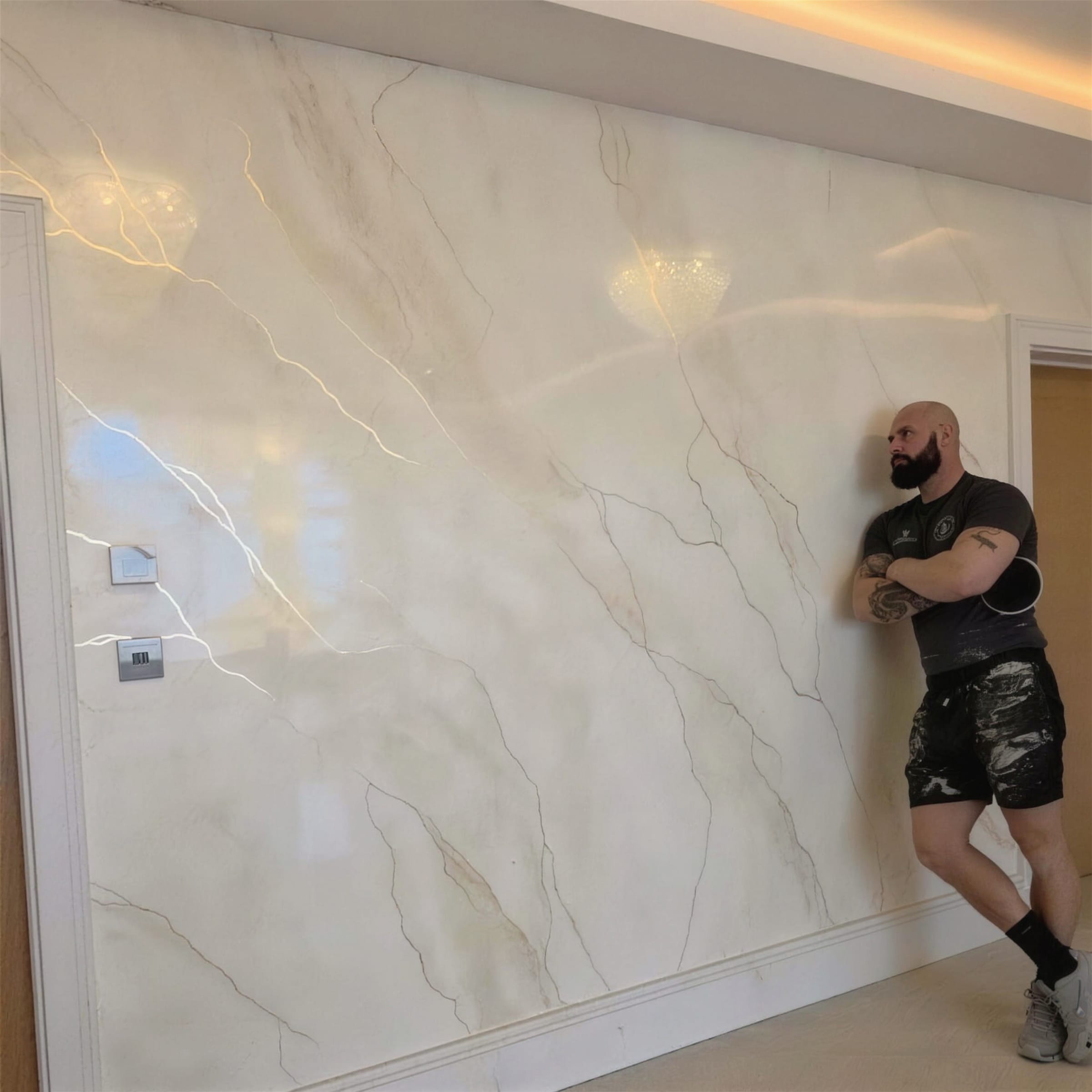 Daros leaning against a full-height polished cream plaster wall veined in gold, warm cove lighting washing the corridor ceiling