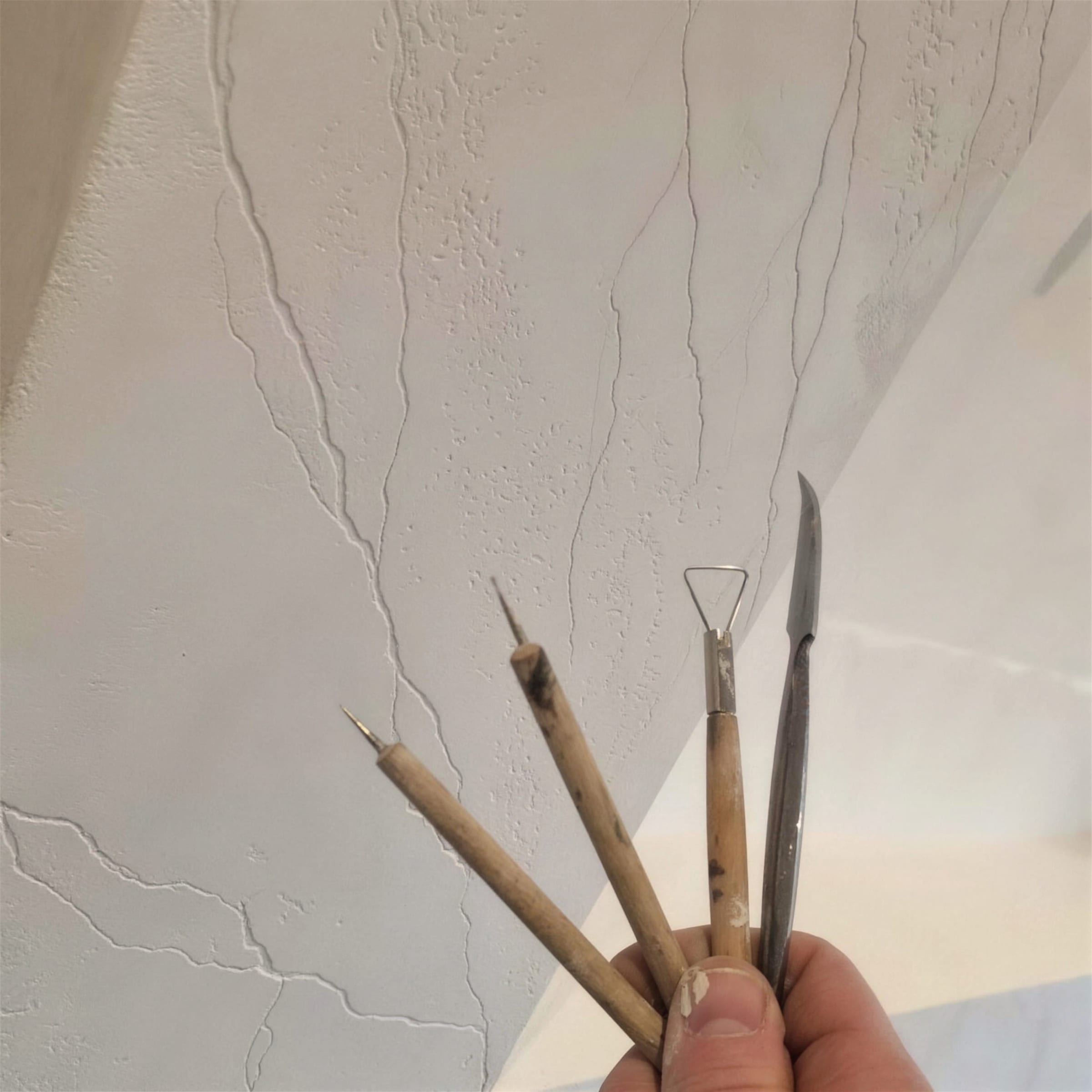 A hand fans four worn vein-drawing tools in front of a white plaster surface scored with fine travertine-style veining.