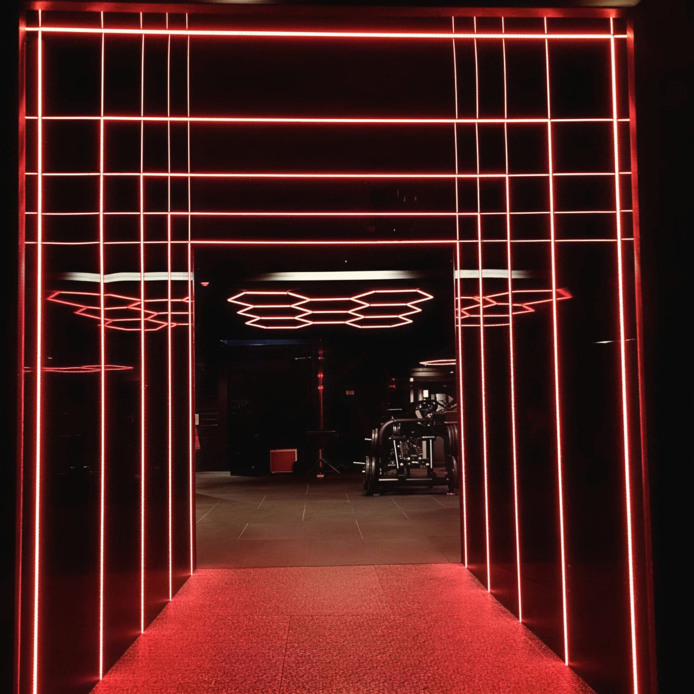 Straight-on view through a red neon-grid LED tunnel toward hexagonal ceiling lights in the gym beyond.