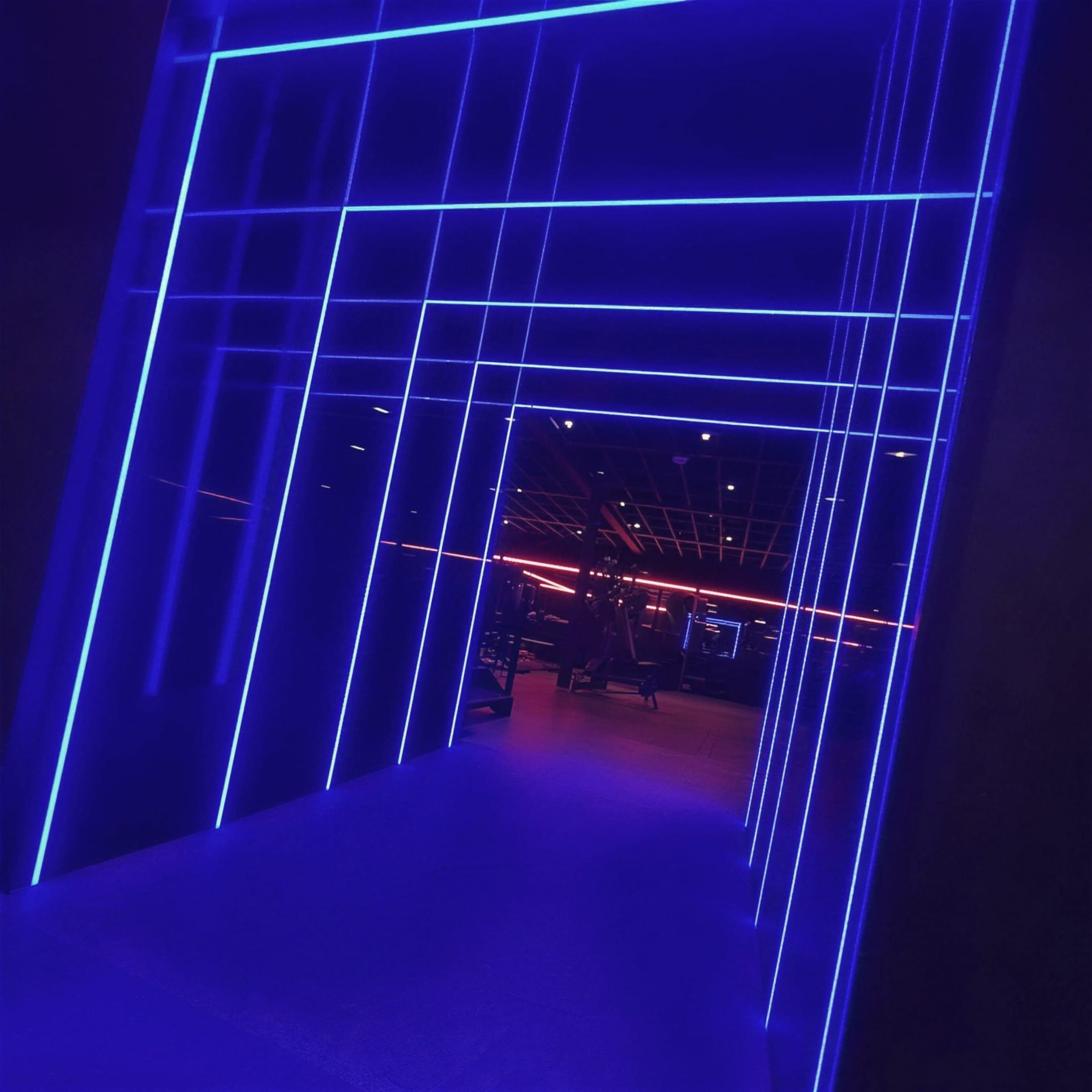 Angled perspective through the electric-blue LED grid tunnel, lines converging toward the gym beyond.