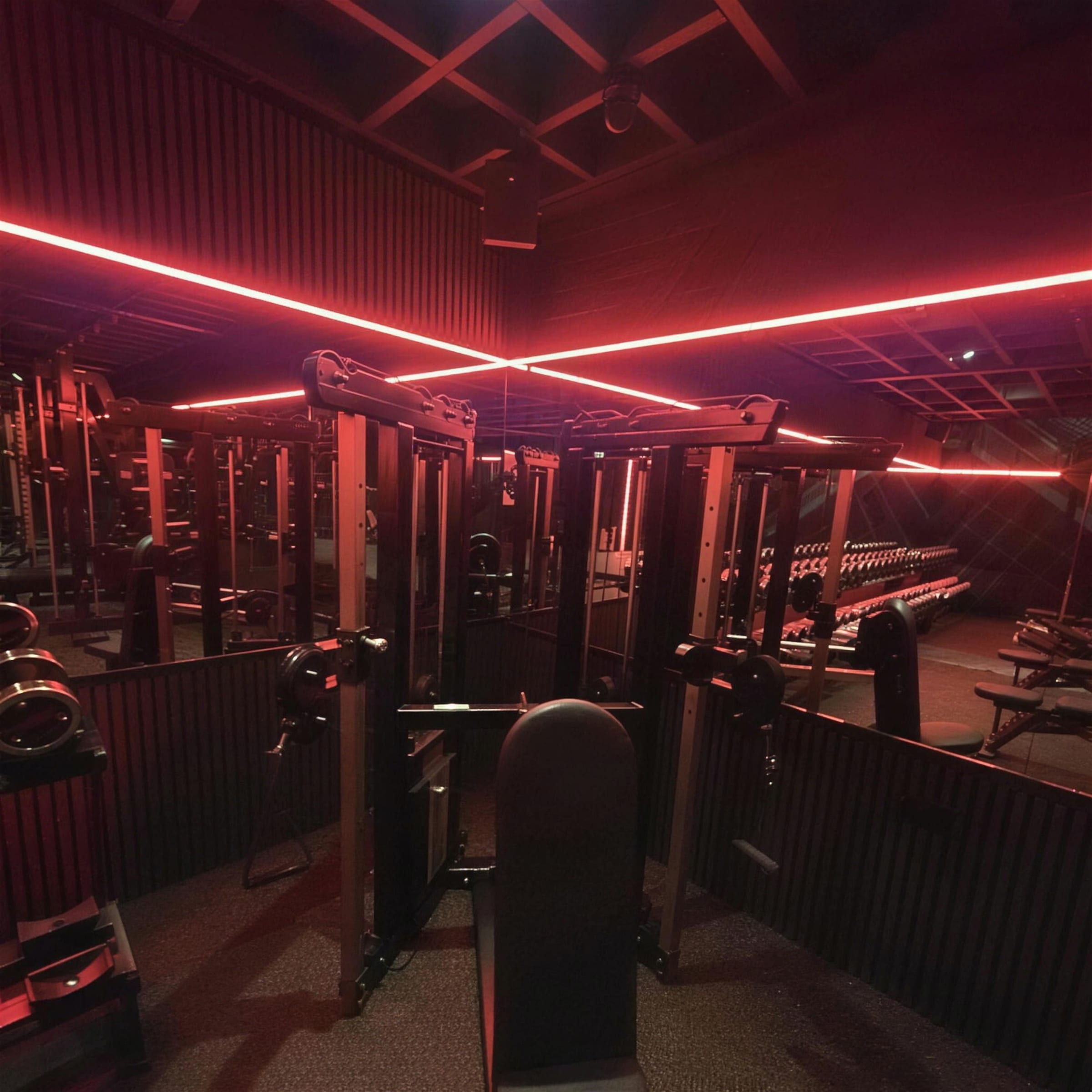 Cable machines and racks under crossing red LED beams in a dark timber-slatted gym interior.