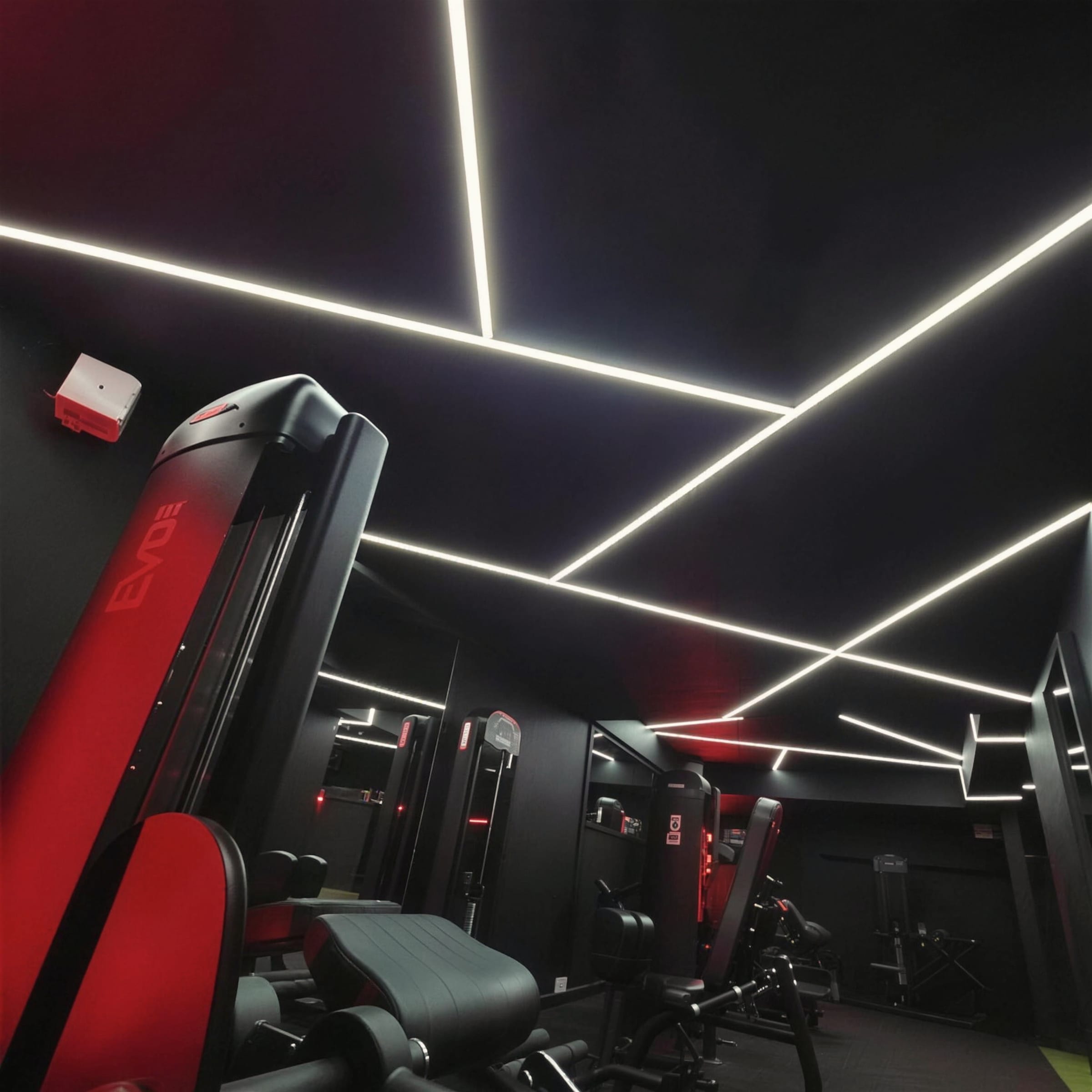 Angular white LED strip lines cutting across a matte black gym ceiling, red-accented equipment below.
