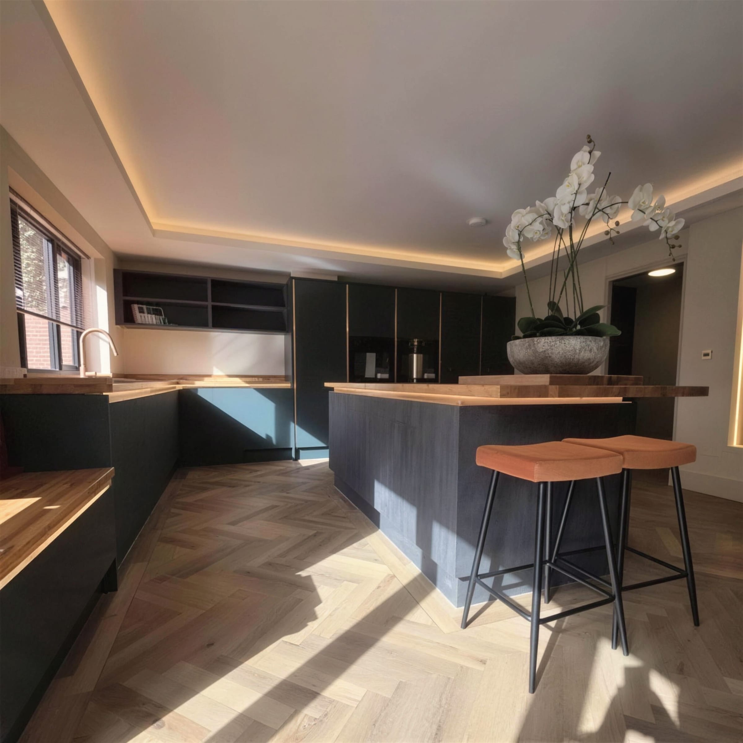 Dark-cabinet kitchen under a warm perimeter LED cove, sunlight raking across herringbone oak flooring, orchids and leather stools at the island.