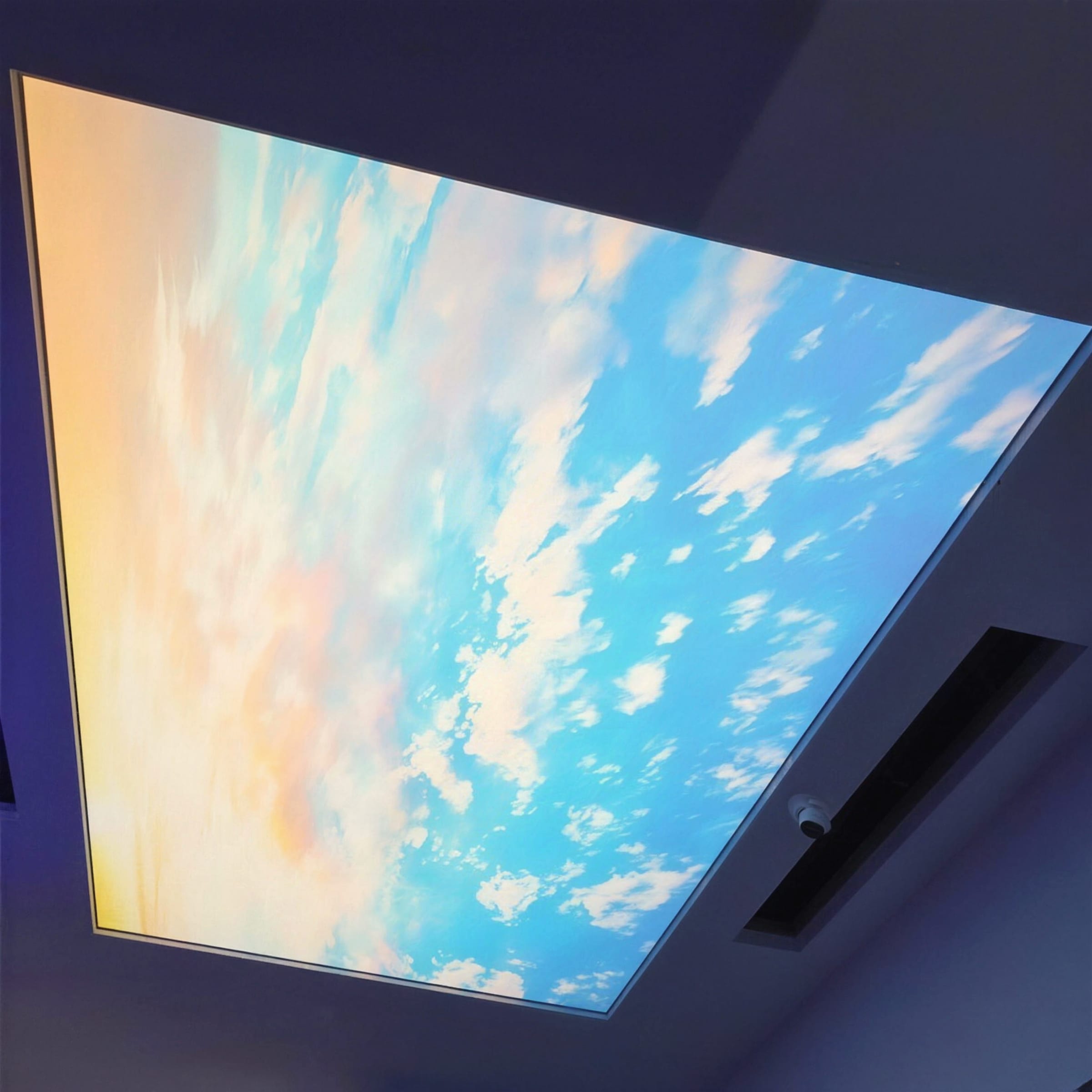 Tight crop of a backlit sky ceiling panel, sunset clouds fading from warm gold into vivid blue with no room clutter in frame.