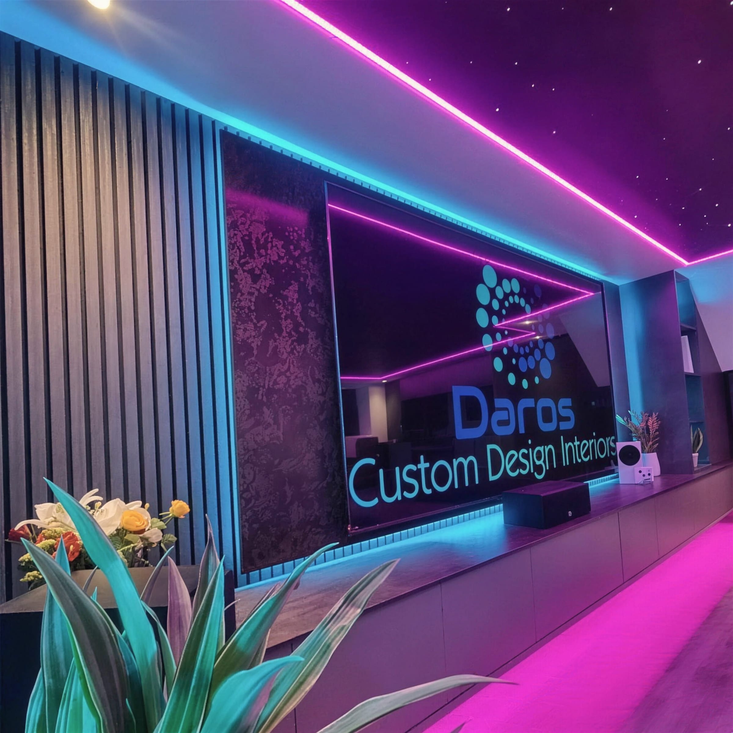 Home-cinema wall wrapped in magenta and blue LED strip lighting with textured black feature panels and planting in the foreground.