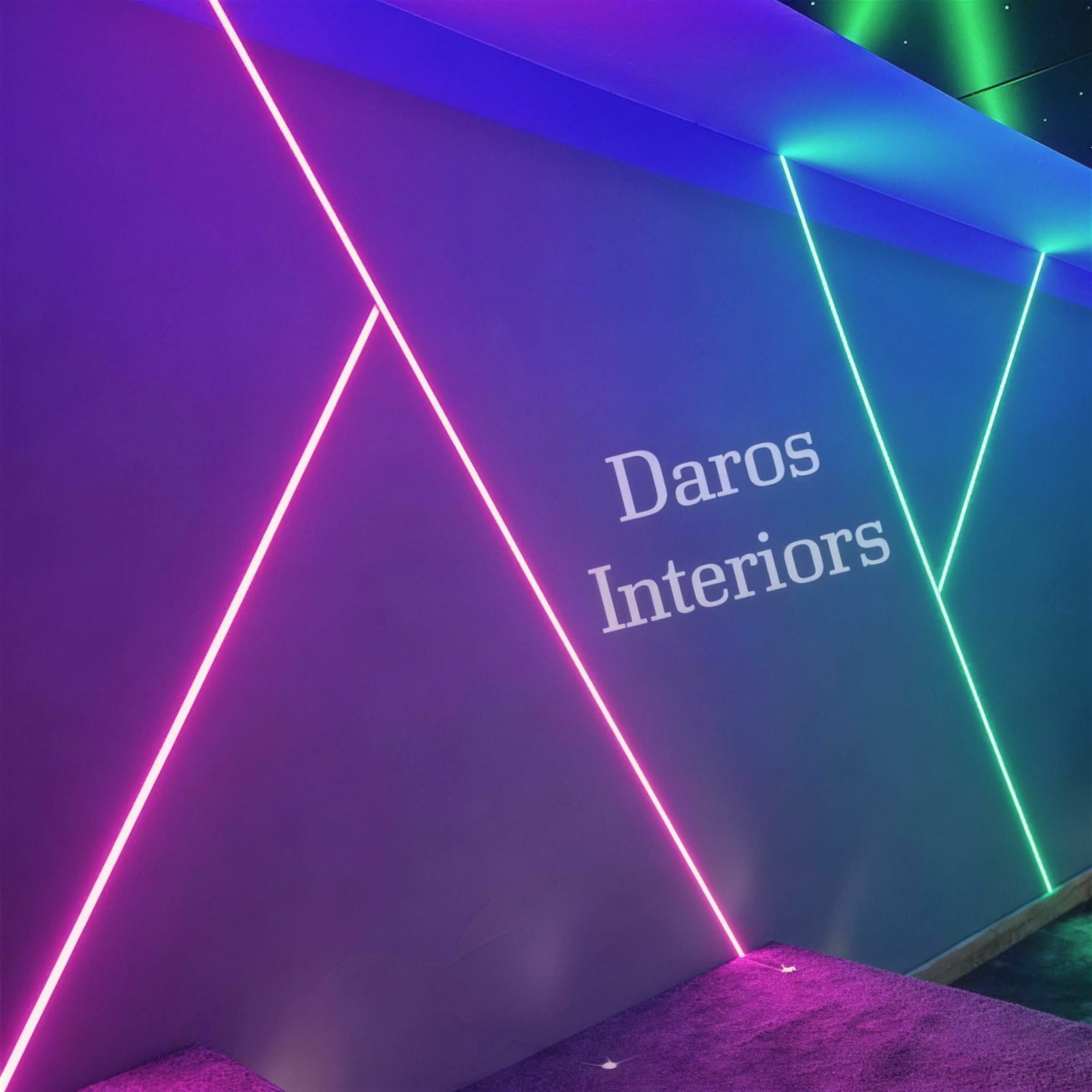 Angled detail of magenta, blue and green neon LED lines forming geometric shapes on a cinema room wall, with a Daros Interiors watermark overlay.