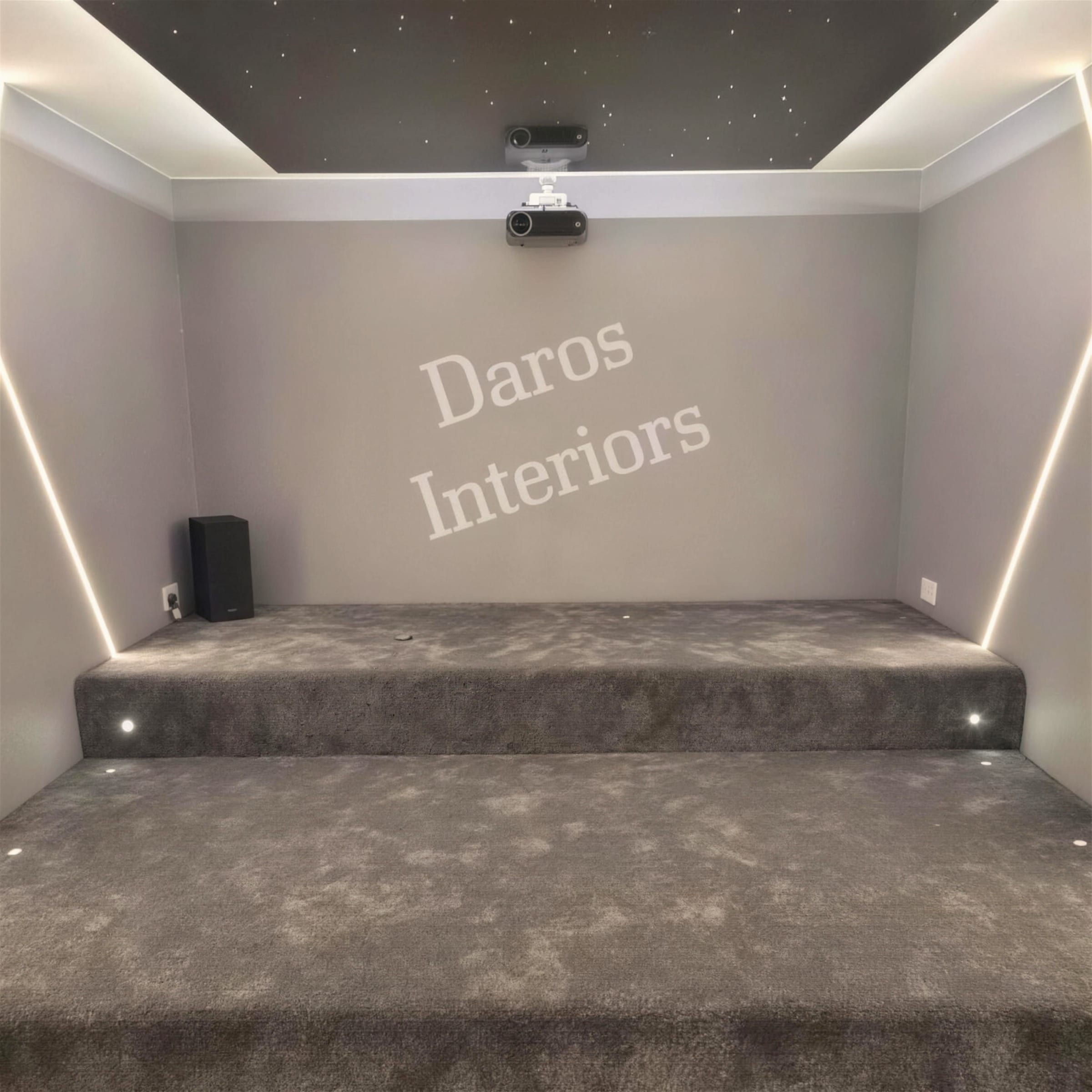 Cinema room with house lights up showing grey carpeted seating tiers, recessed LED cove and wall-wash strips, and a dark star-ceiling panel above.