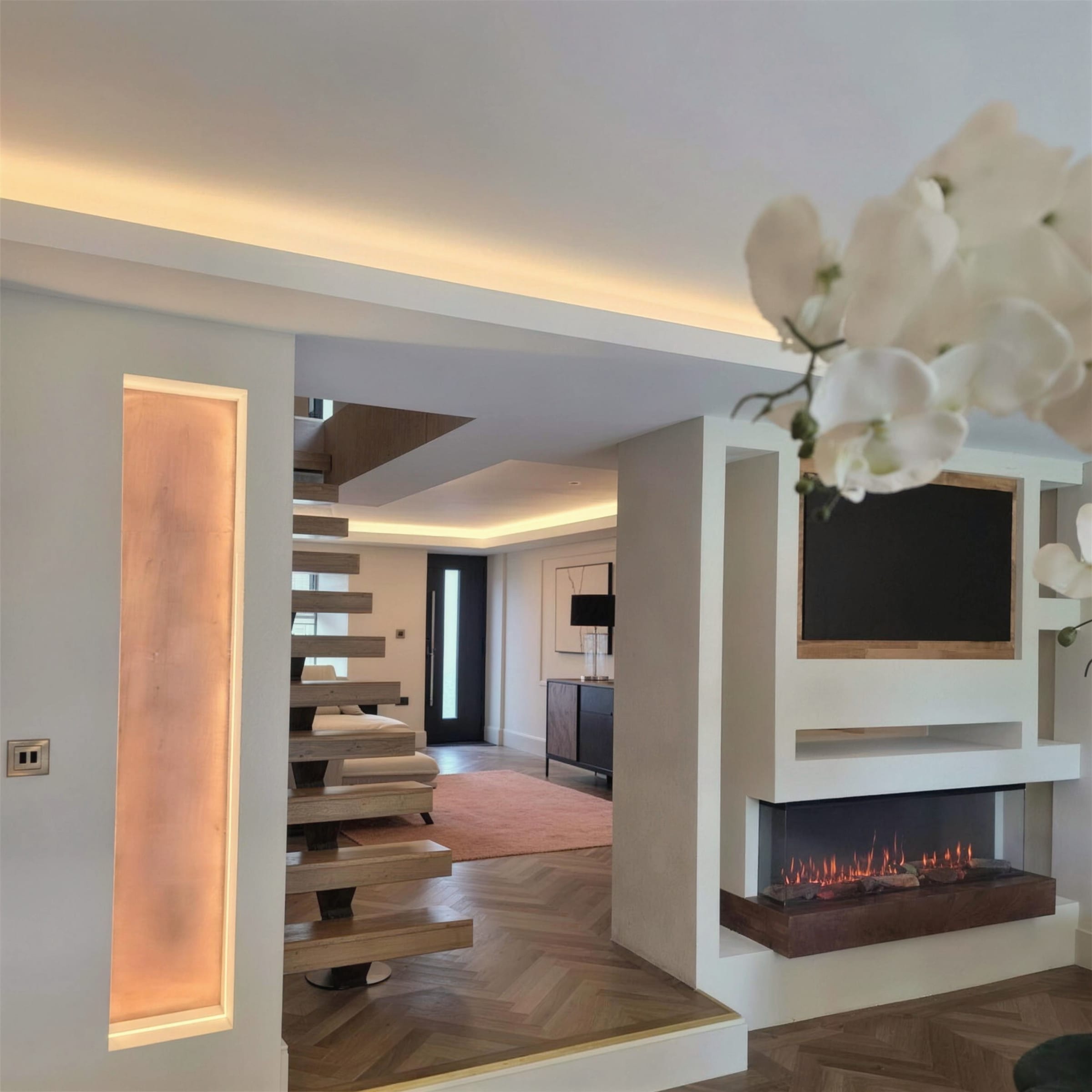 Luxury hallway with an amber backlit alabaster-effect panel, floating oak stairs, warm LED cove ceiling and a plaster fireplace media wall, orchid in the foreground.