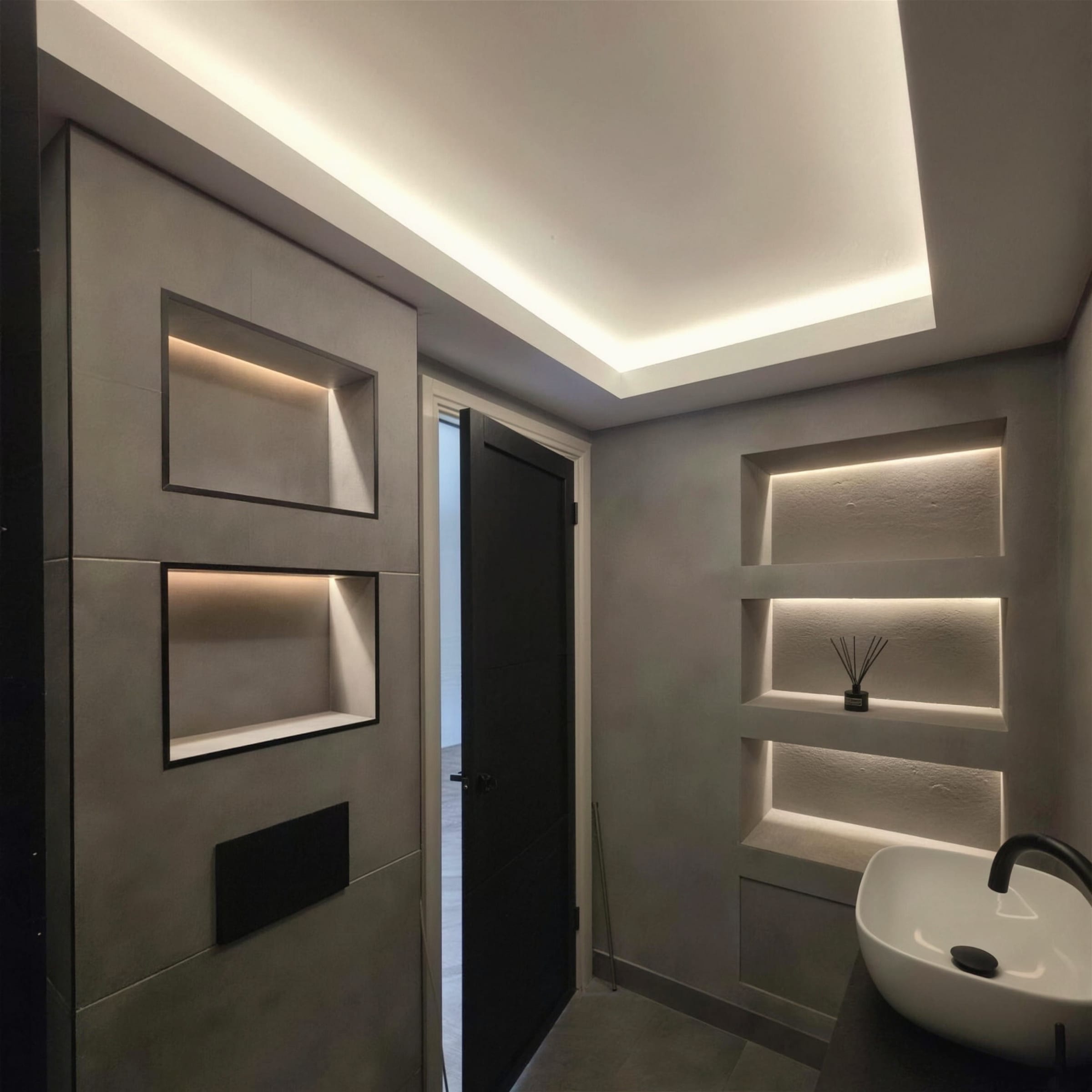 Moody charcoal microcement bathroom with warm LED-lit storage niches, cove-lit ceiling, black-framed shower screen and white vessel basin.