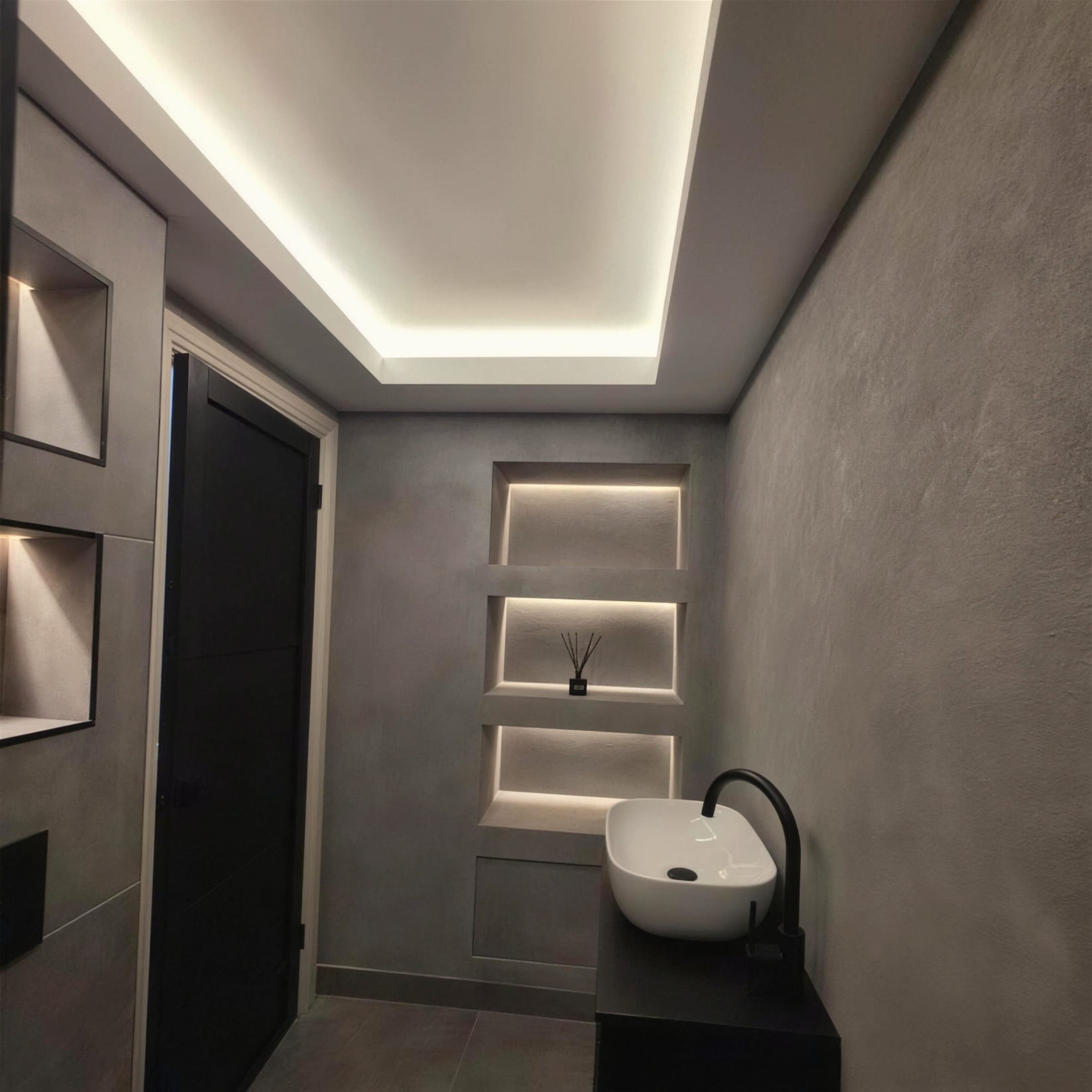 Seamless grey microcement walls and niches under a crisp LED tray ceiling, with a white countertop basin and matt-black tap; clean, moody and sharp.