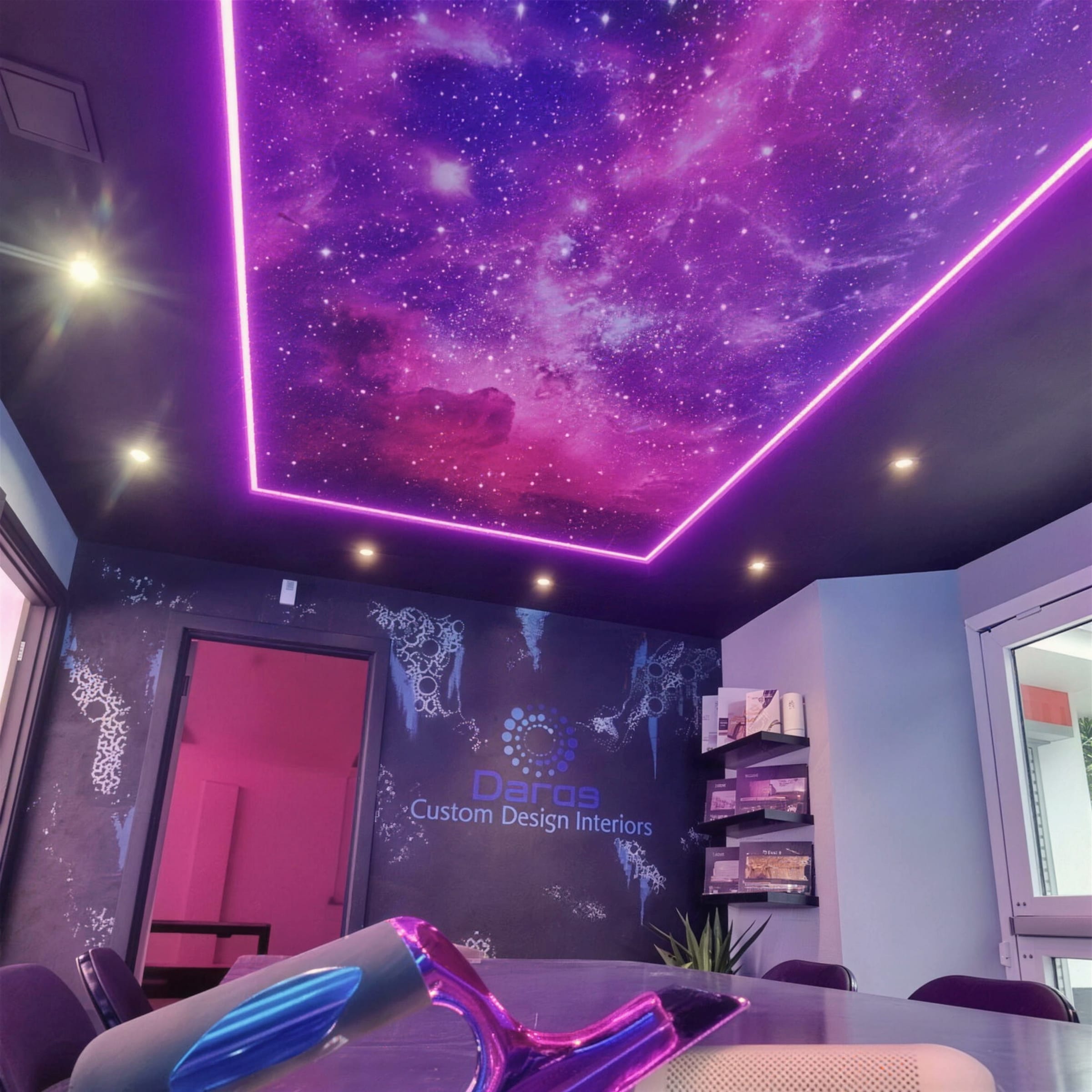 Printed nebula ceiling panel with pinpoint stars, framed by a violet neon perimeter and downlights in the showroom boardroom.