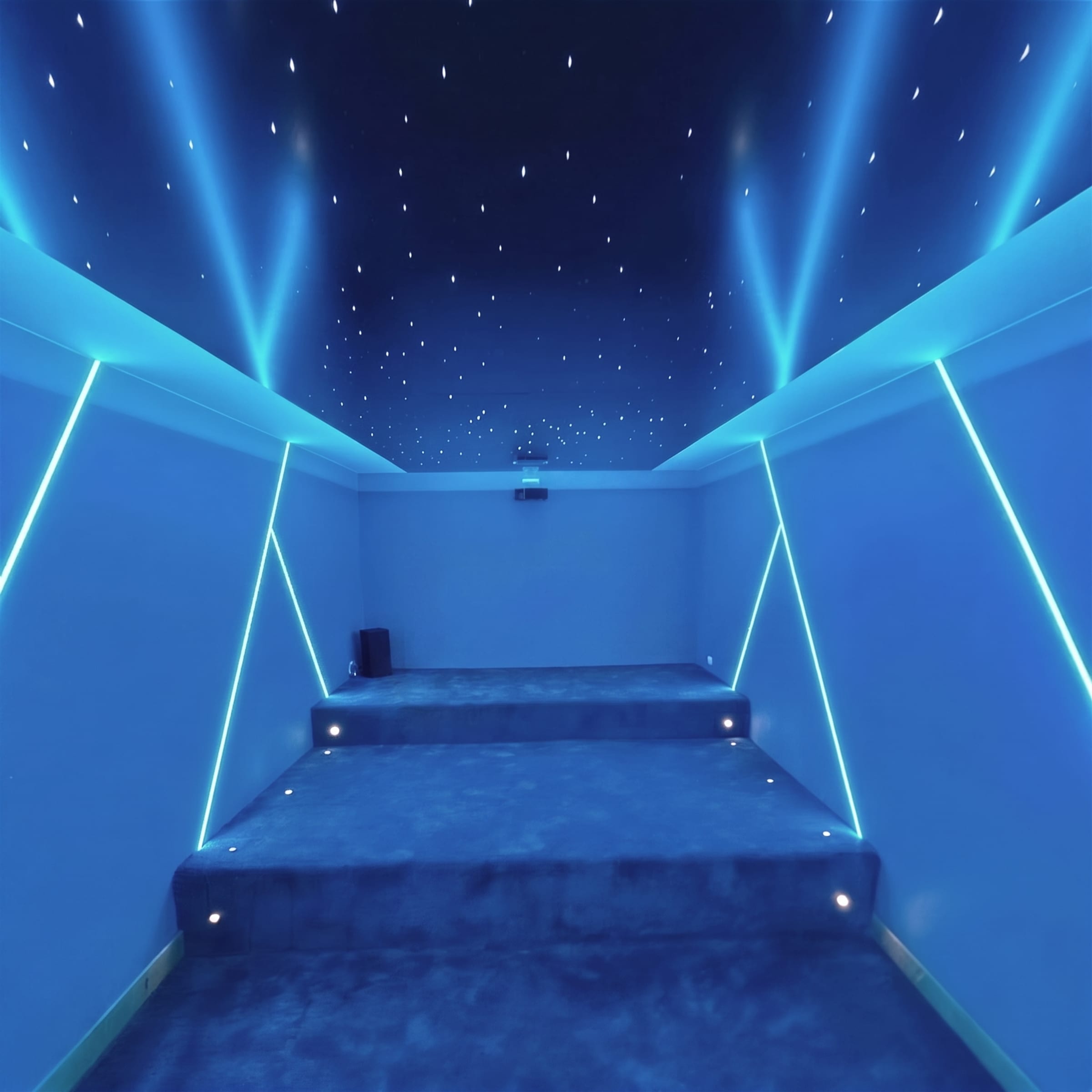 Symmetrical view up a blue-carpeted cinema aisle toward a star-field ceiling with sweeping blue light beams and LED-edged walls, watermarked Daros Interiors.