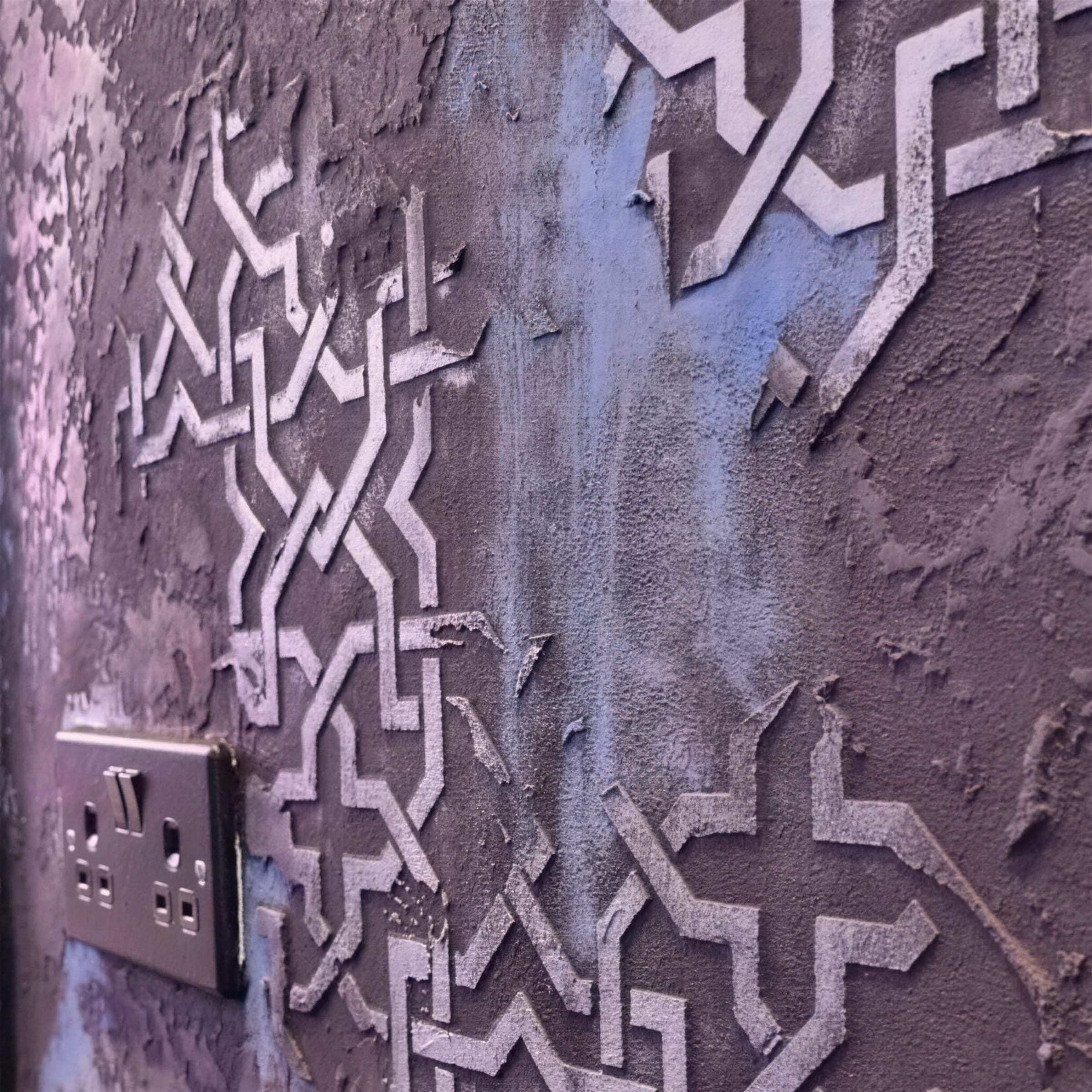 Close view of a plum-and-blue distressed plaster wall with a raised Moorish star-lattice stencil, a chrome double socket set into the finish.