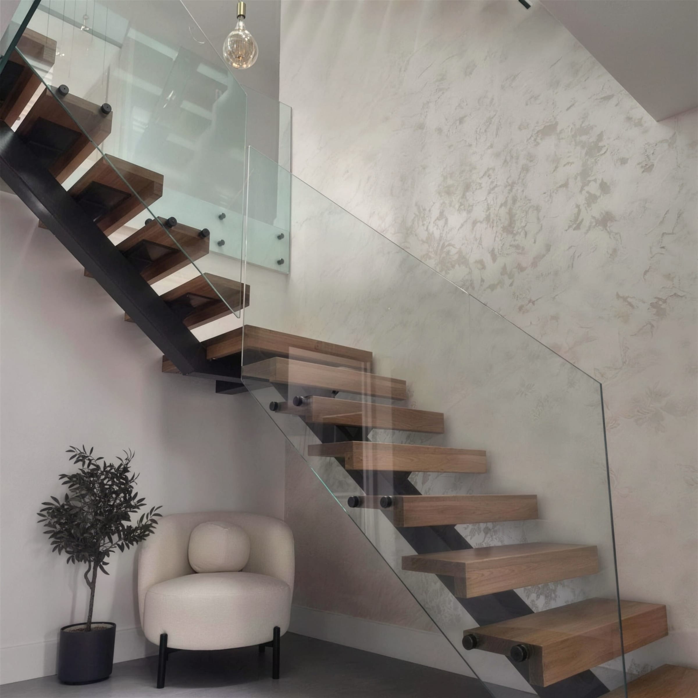 Floating oak staircase with glass balustrade against a double-height pearlescent marbled plaster wall, styled with an olive tree and boucle chair.