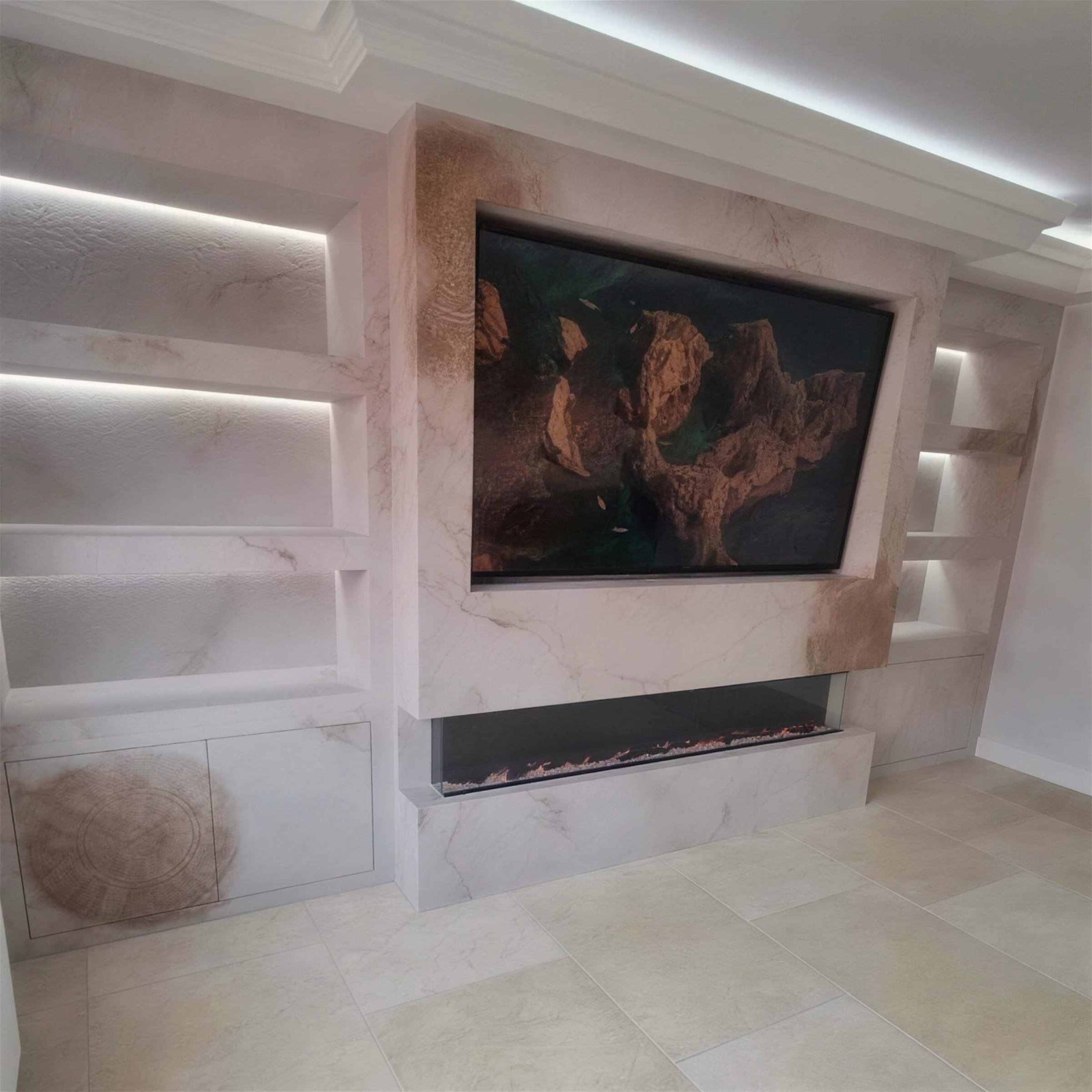 Full media wall in cream travertine-effect plaster with LED-backlit display shelves, recessed TV and linear fire, over travertine flooring.
