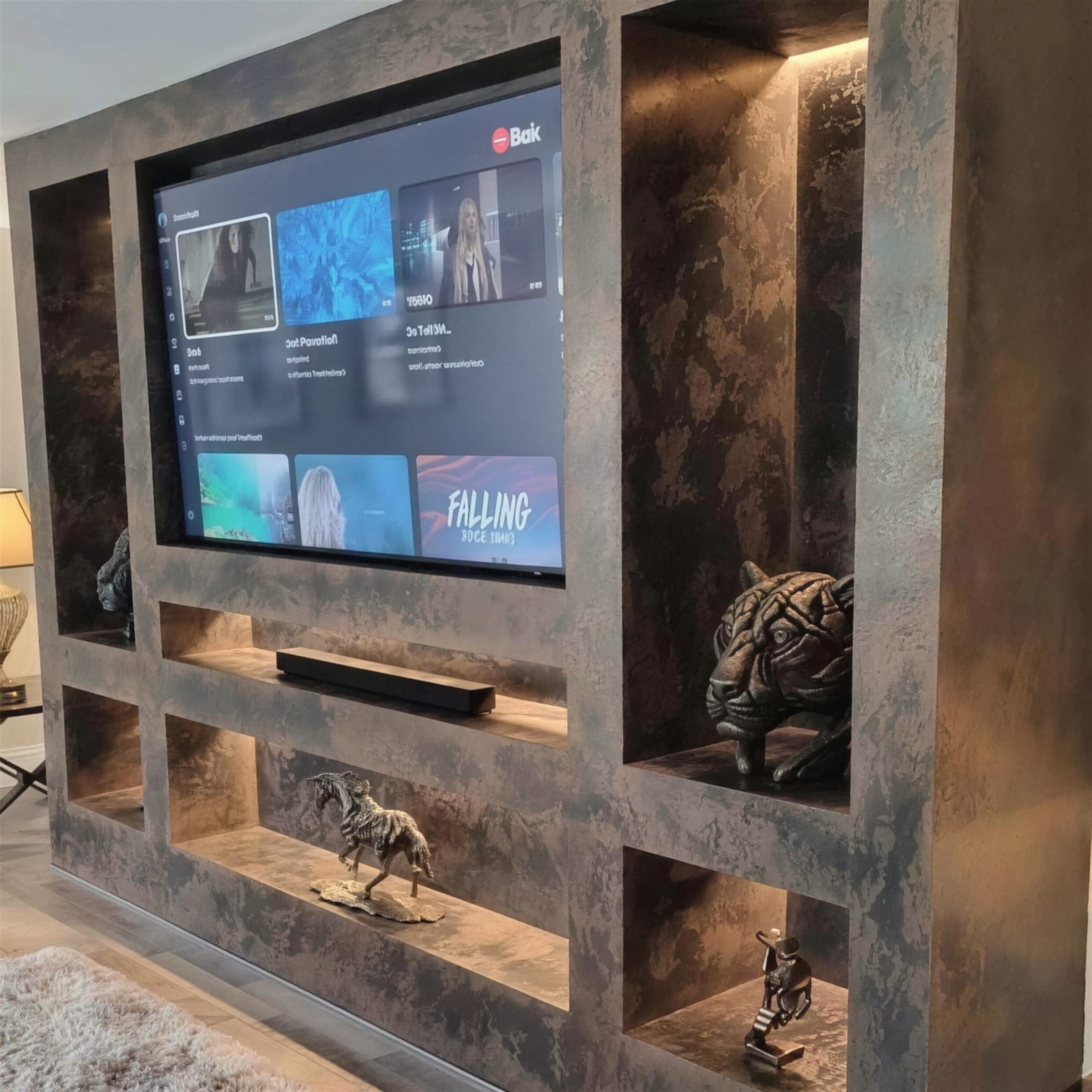 Dark aged-bronze metallic plaster media unit with warm-lit niches holding bronze sculptures around a wall-mounted TV.
