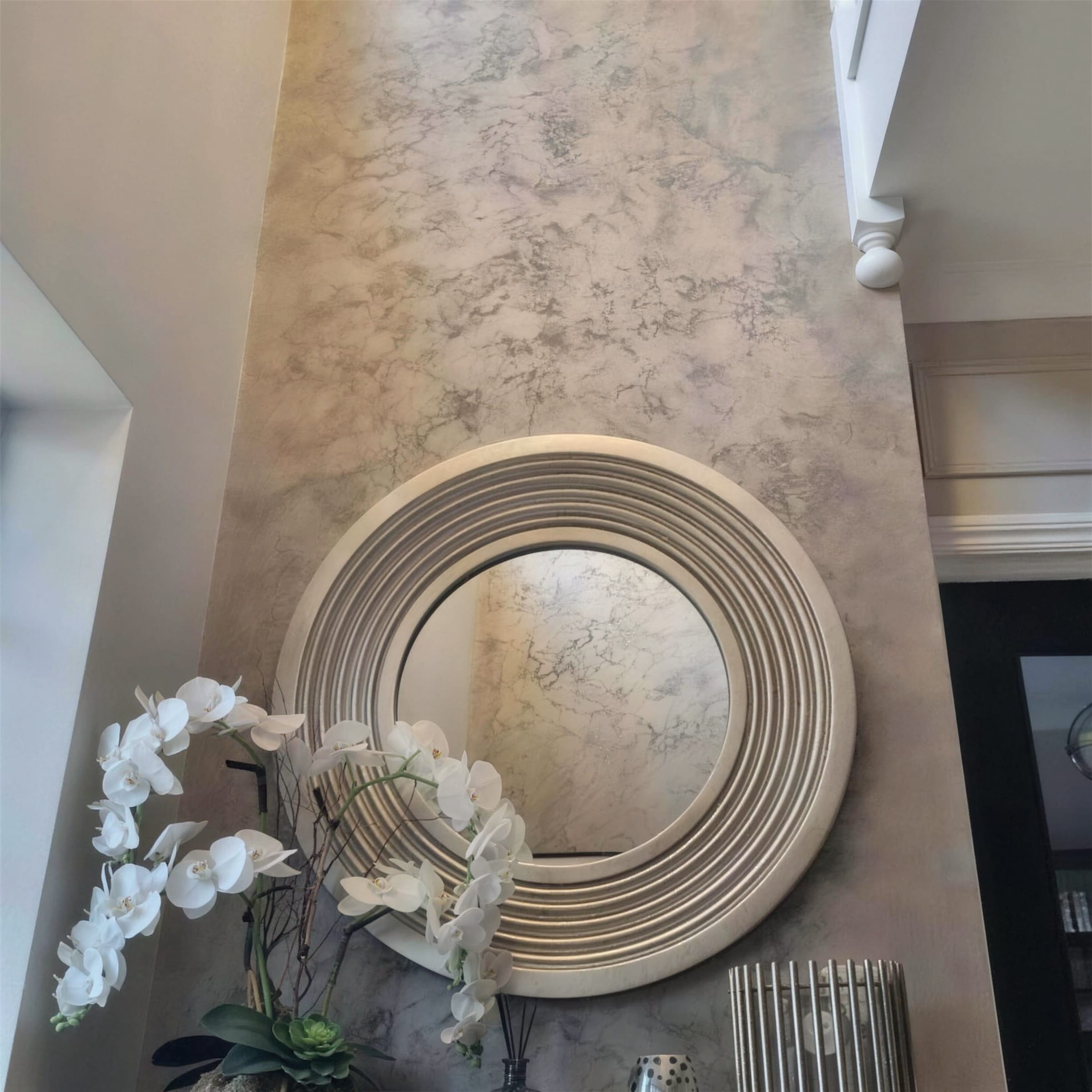 Double-height champagne marbled plaster wall with a ribbed round mirror and white orchids in the foreground.