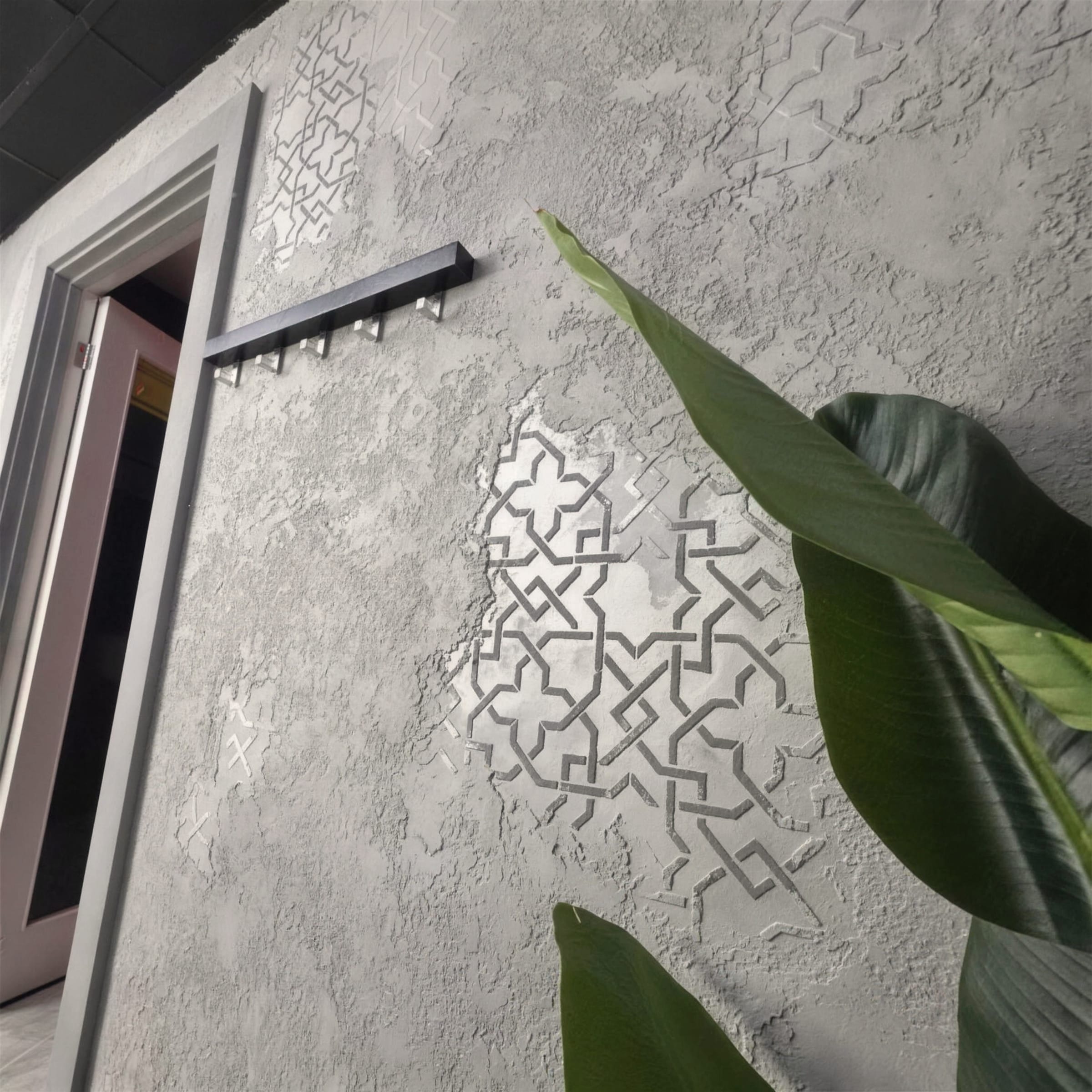 Angled view up a heavily textured grey plaster wall with silver stencil medallions, black coat rail and doorway at left.