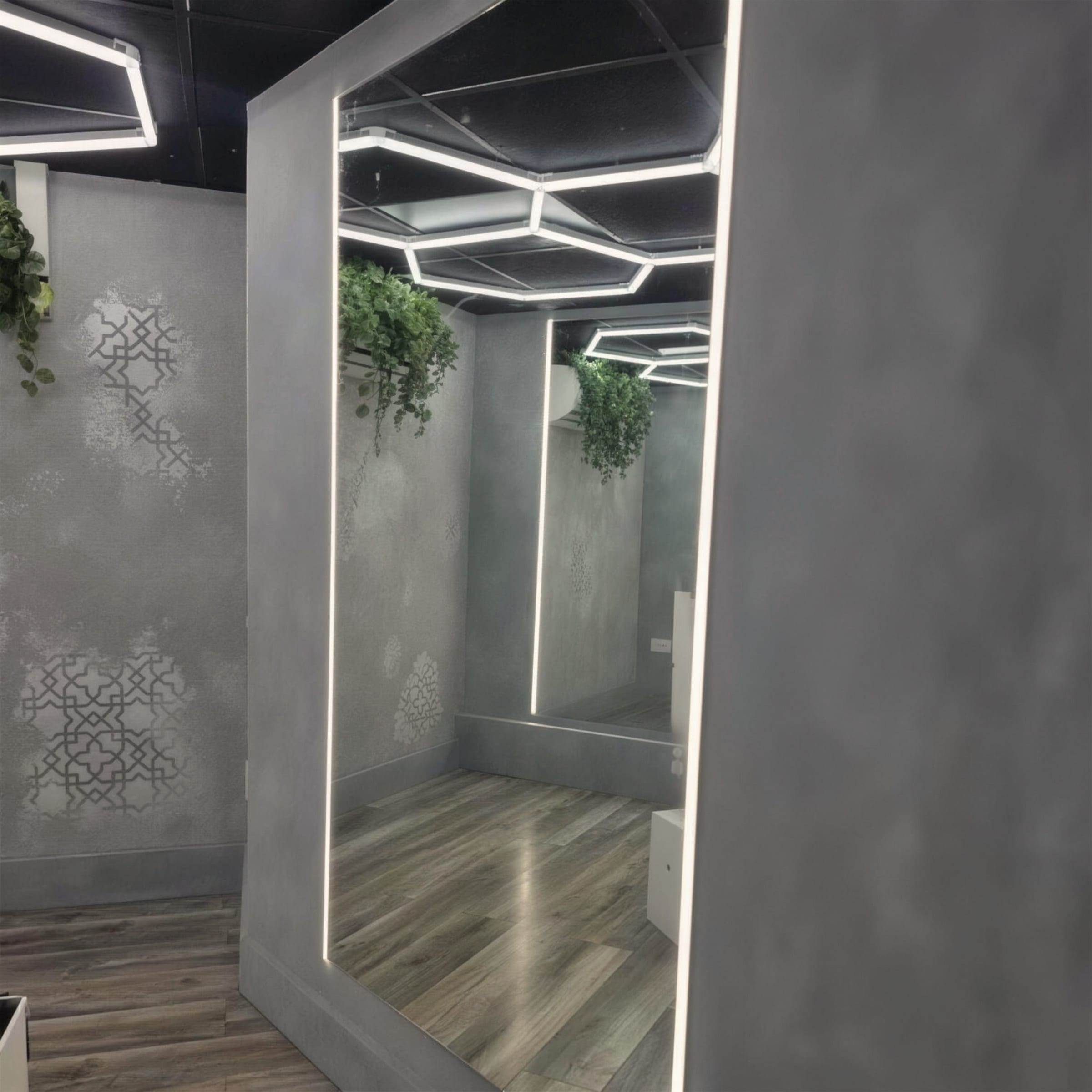 Grey plaster-clad mirror column with LED edge lighting reflecting hexagon ceiling lights and trailing greenery.