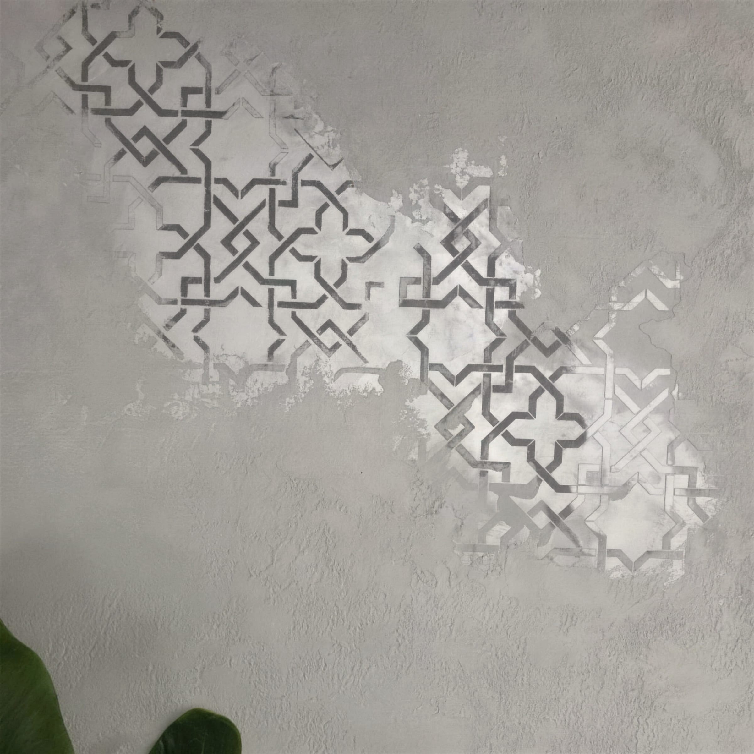 Flat frontal detail of silver metallic geometric stencil pattern emerging from soft grey textured plaster, leaf at the bottom edge.