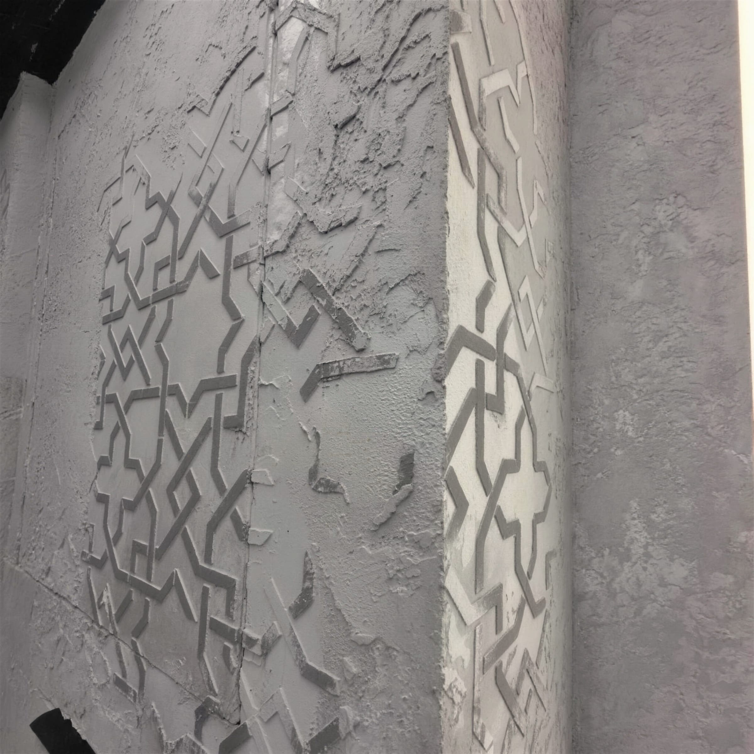 Close detail of a grey heavy-texture plaster column with carved interlocking star-and-cross geometric relief wrapping the corner.