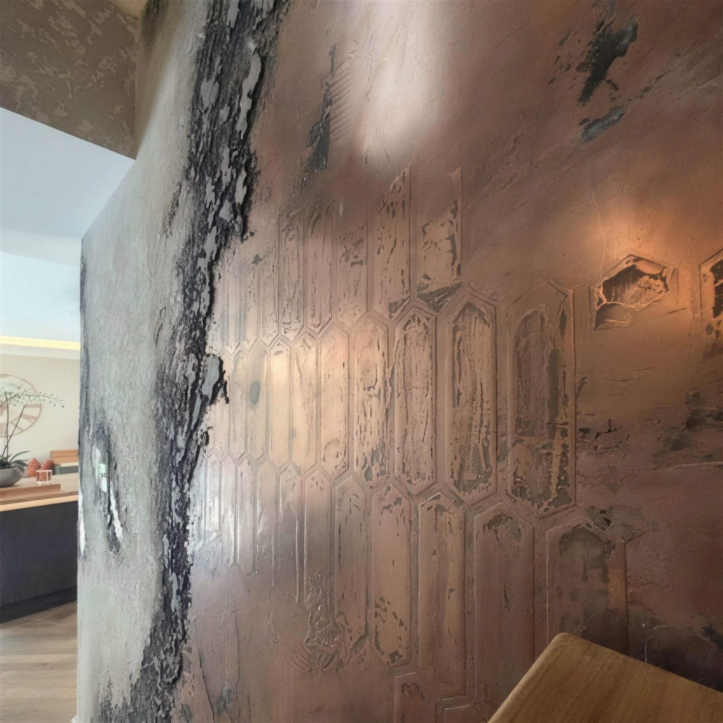Burnished copper metallic plaster with charcoal oxidised patination and an embossed hexagonal motif, kitchen glimpsed beyond the wall edge.