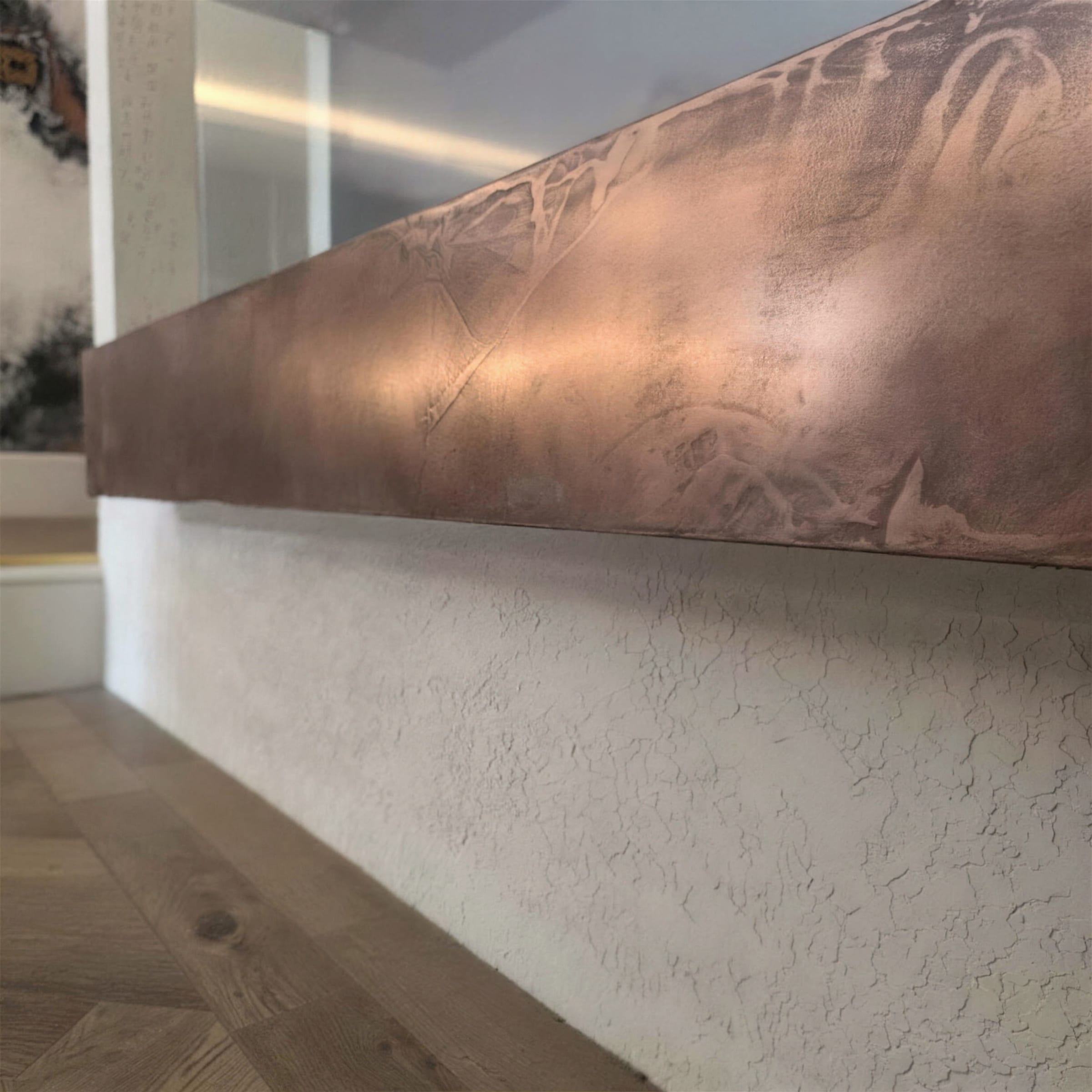 Close-up of a burnished copper metallic plaster beam over a white rustic-texture plinth, herringbone floor receding below.