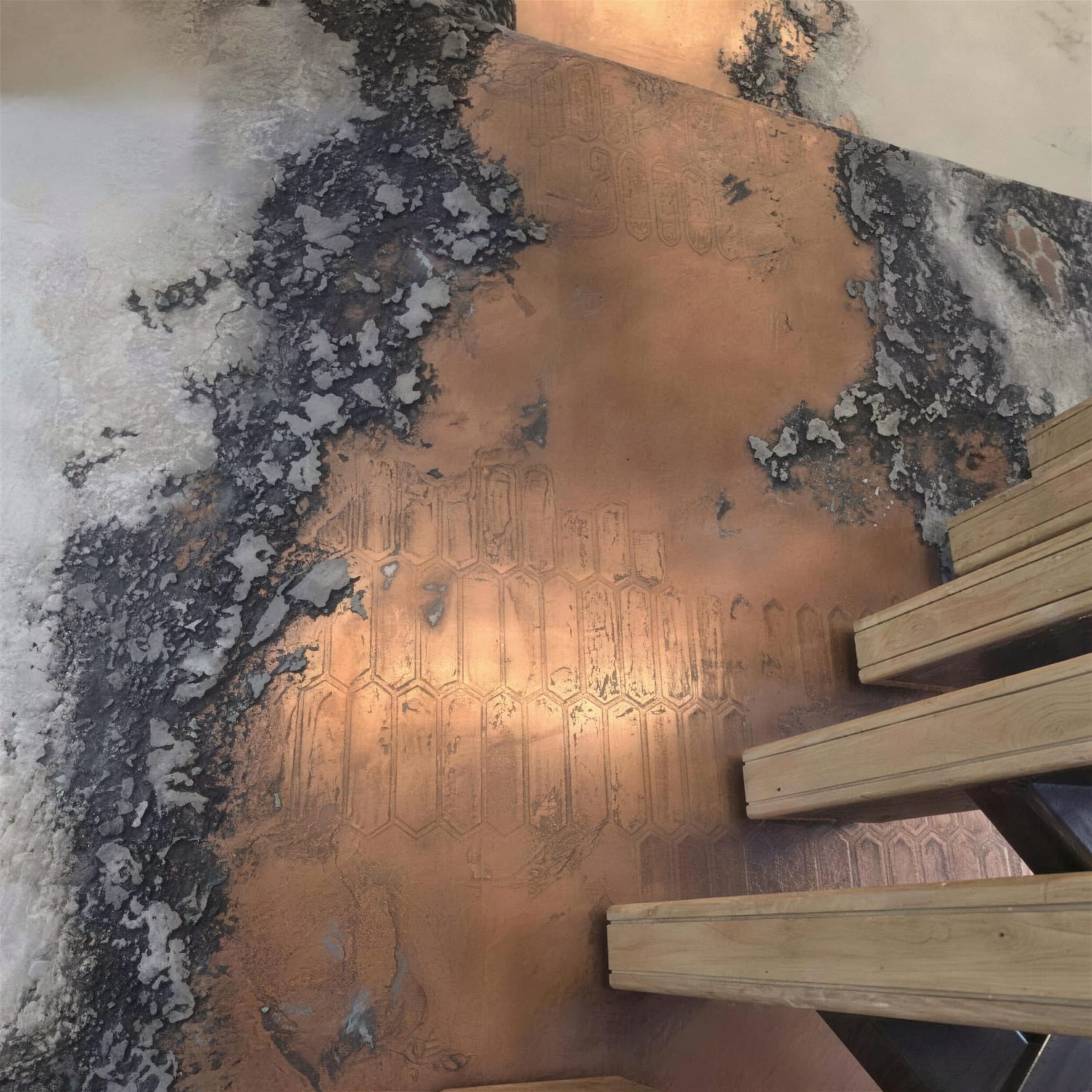 Top-down view along timber stair treads as the embossed copper and charcoal oxidised plaster wall flows down the stairwell.
