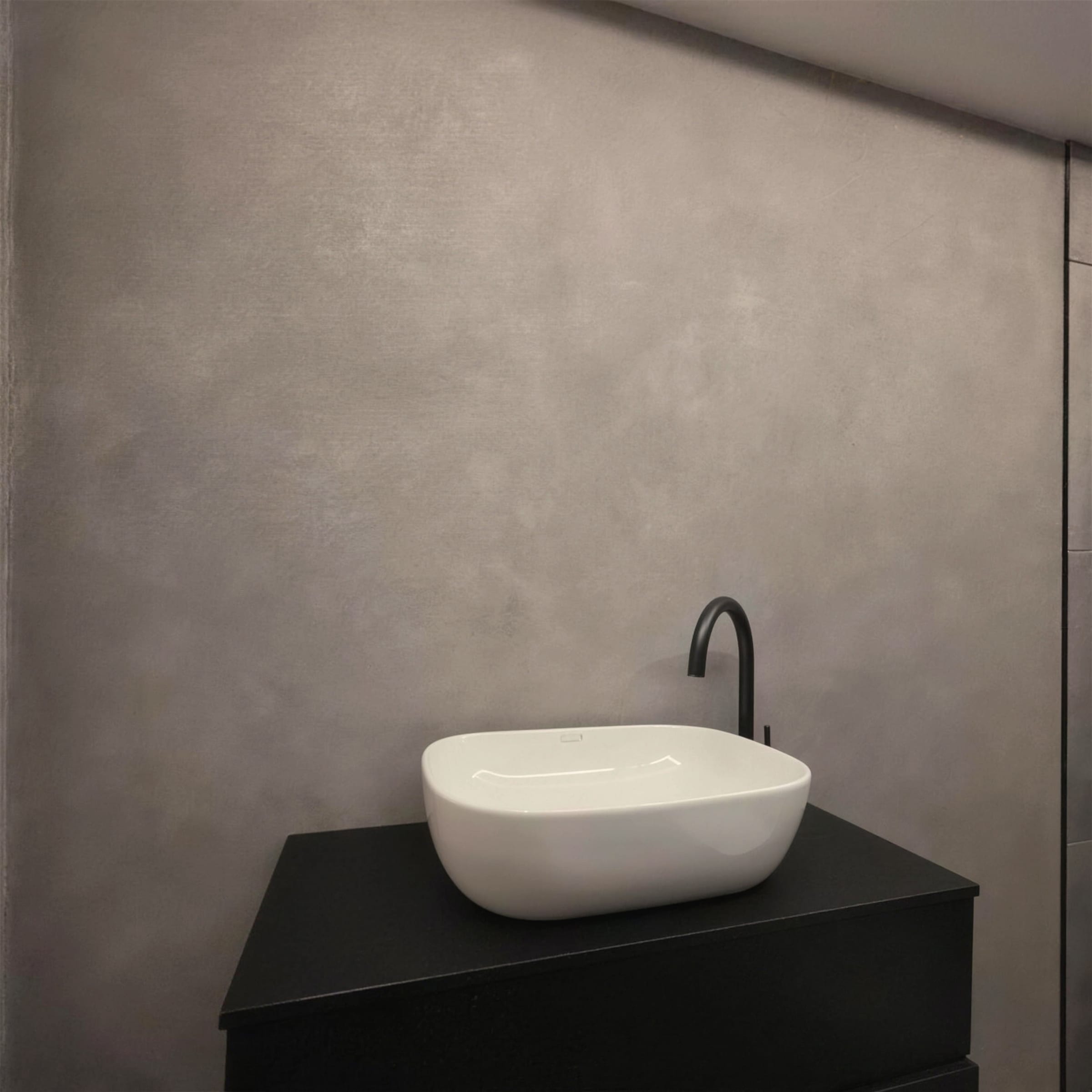 Minimal composition of a cloudy grey polished marmorino wall behind a matte black vanity, white basin and slim black tap.