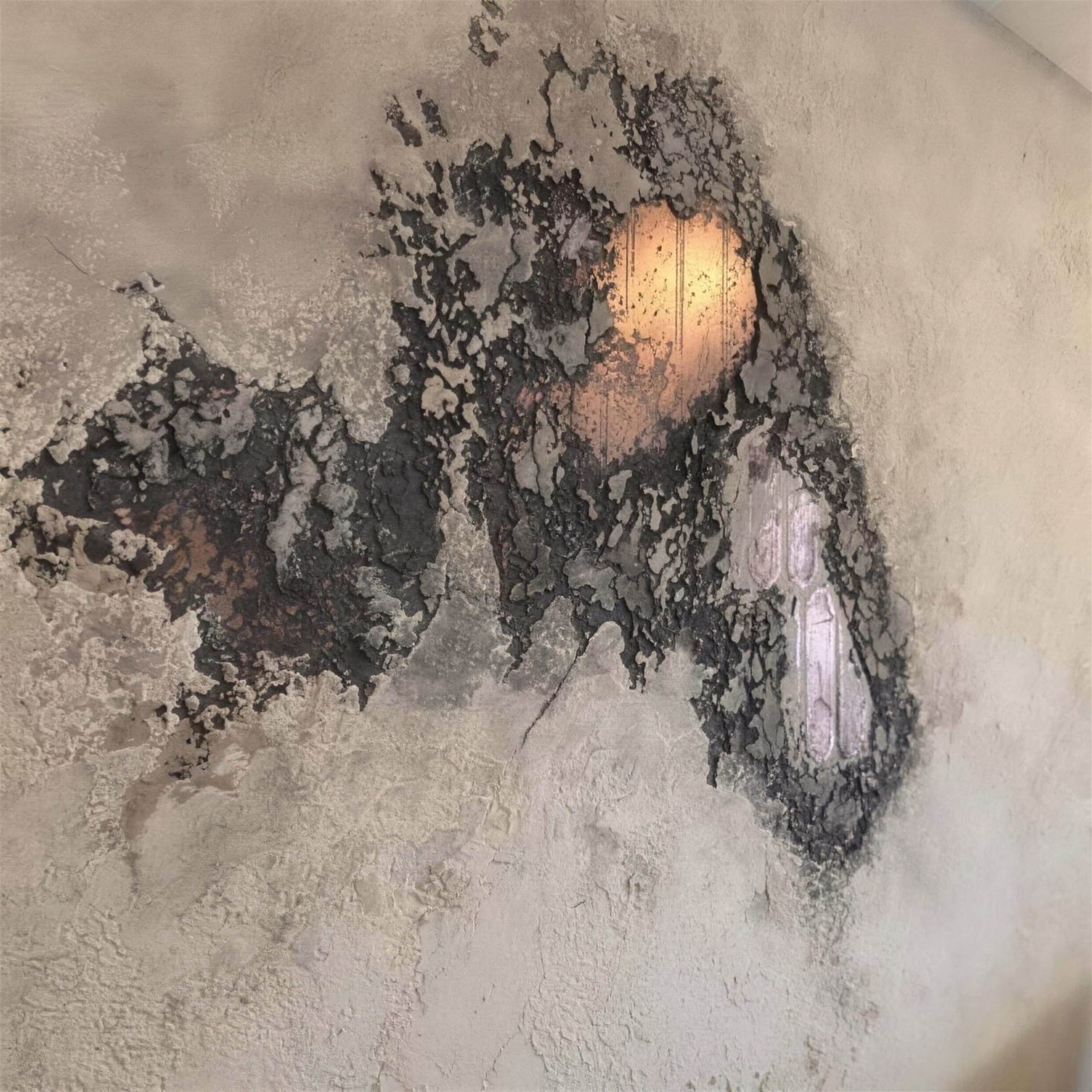Cream rustic travertine wall with a dark oxidised copper patch through which a warm wall light glows.