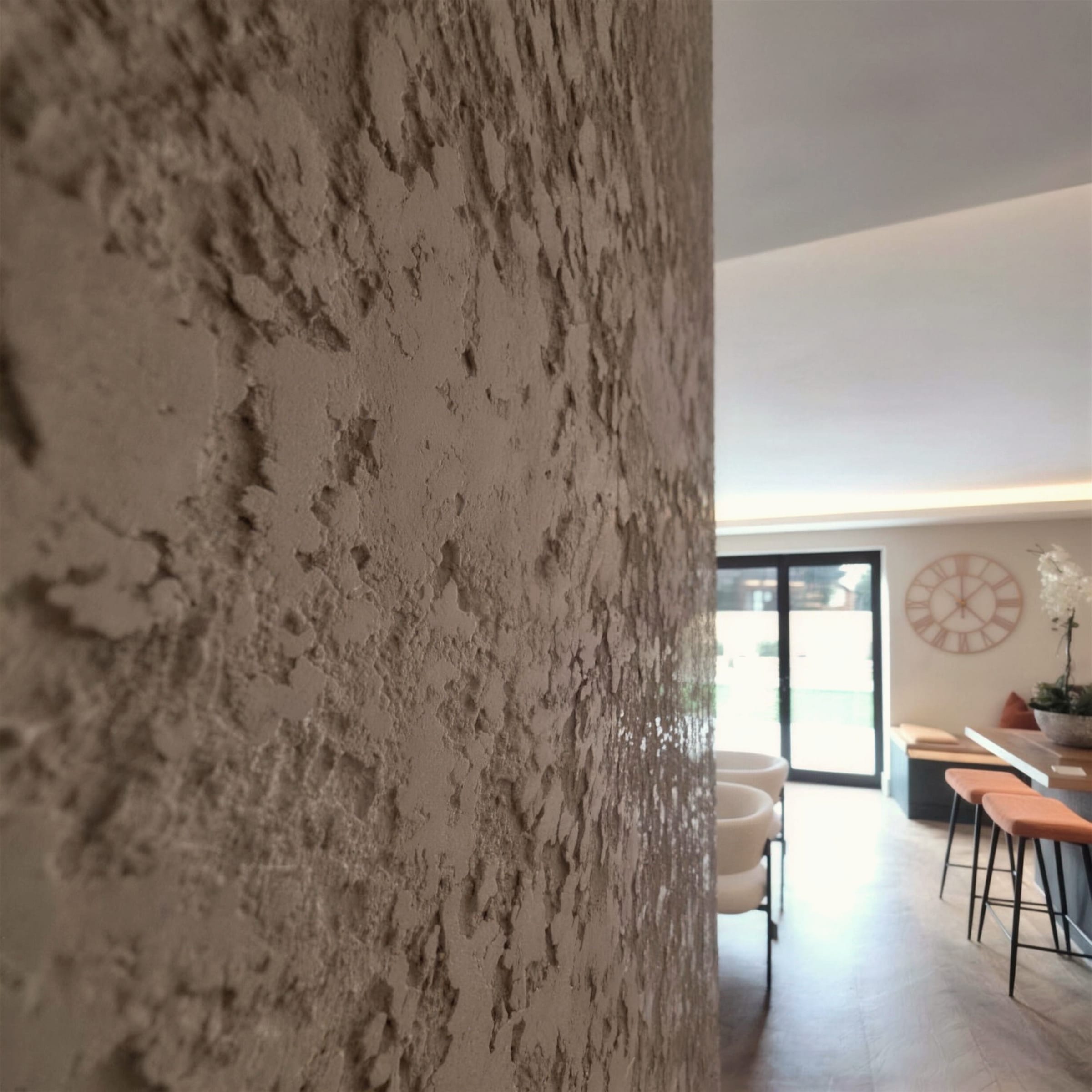 Raking close-up of a heavily textured cream travertine-style plaster wall with the finished open-plan living space softly blurred behind.