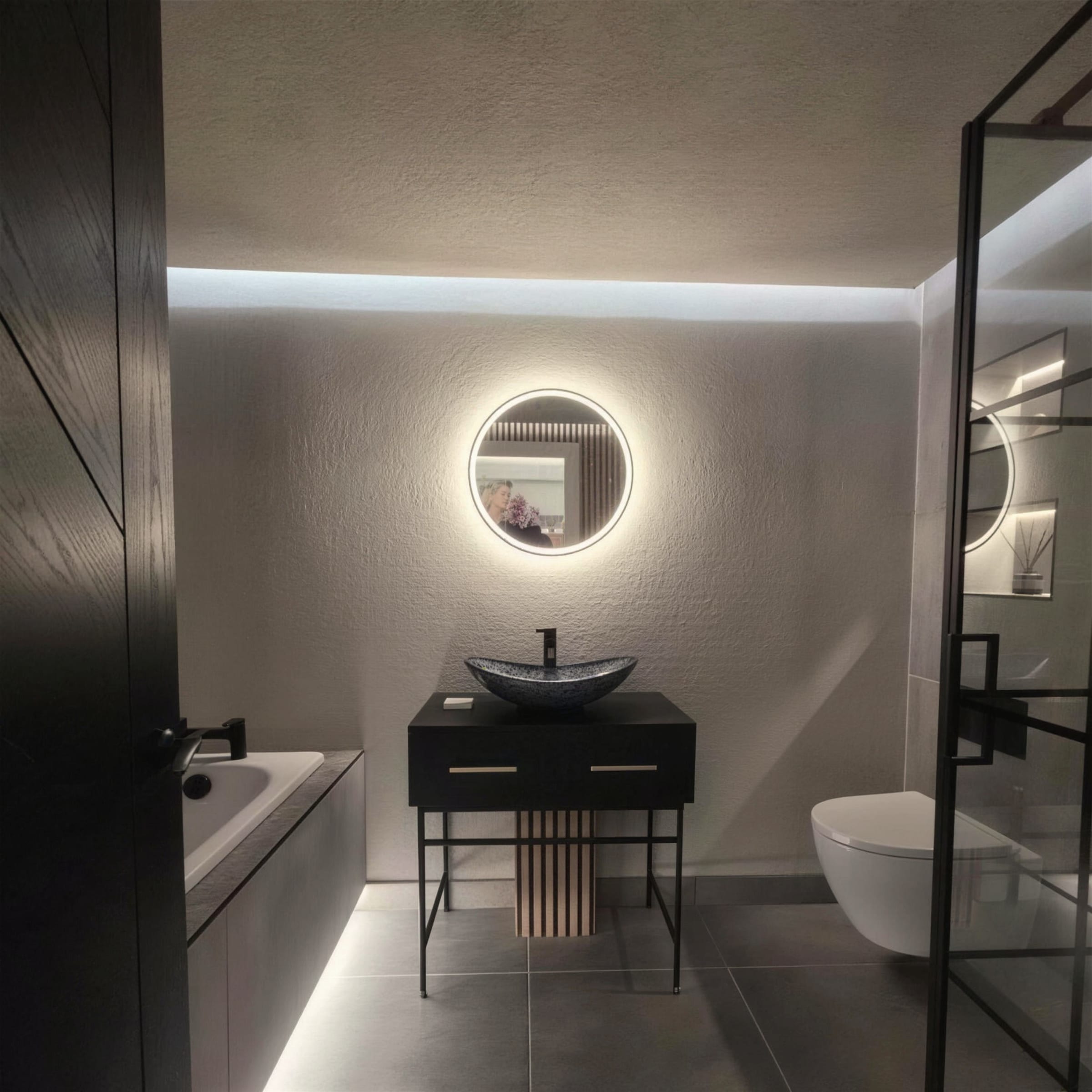Grey textured-plaster bathroom with halo-lit round mirror, stone vessel basin on a black washstand and concealed LED strip washing the ceiling line.