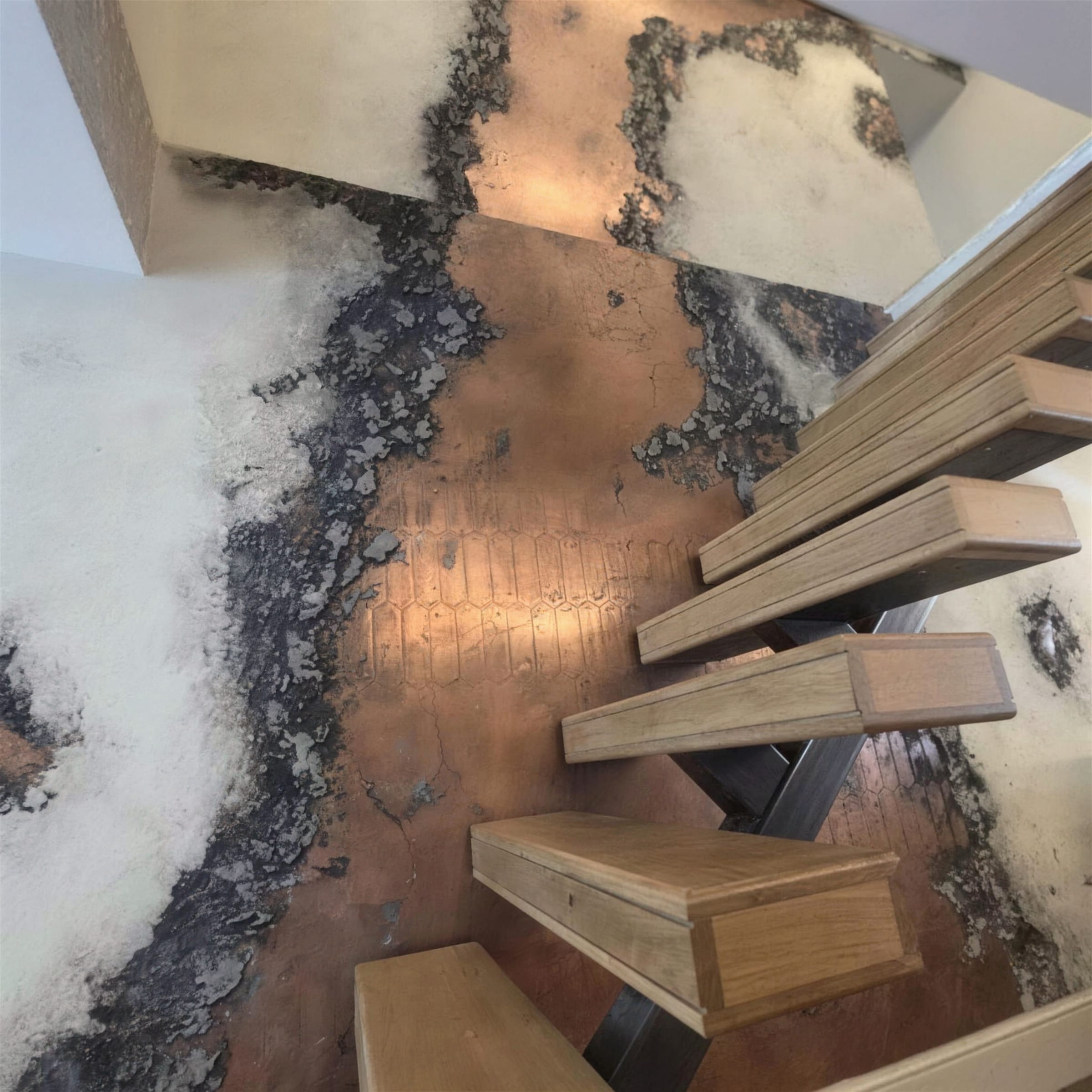 Top-down view over floating oak treads onto a sweeping oxidised copper and black metallic plaster wall running down the stairwell.