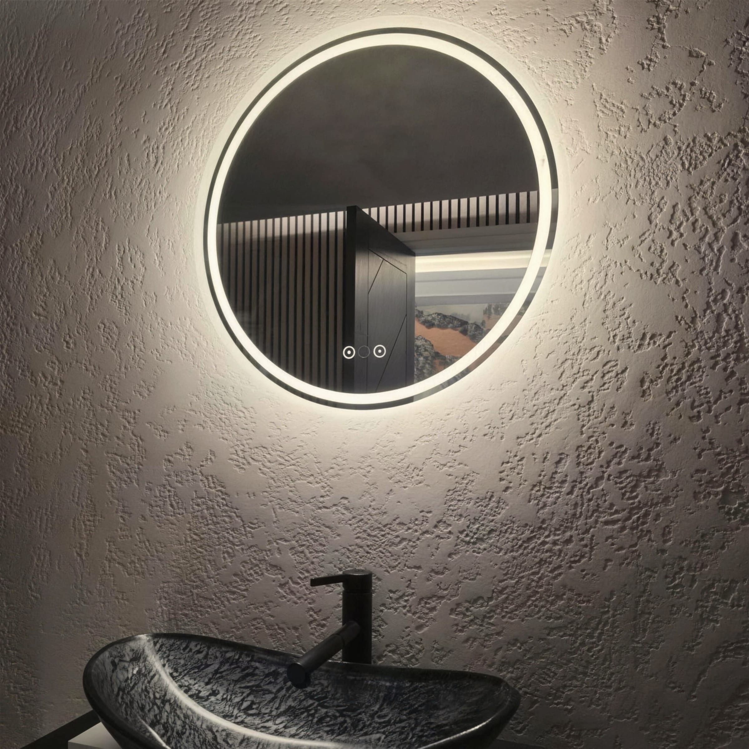 Halo-lit round mirror glowing against a dark stone-textured plaster wall, with a carved stone vessel basin and matt black tap below.