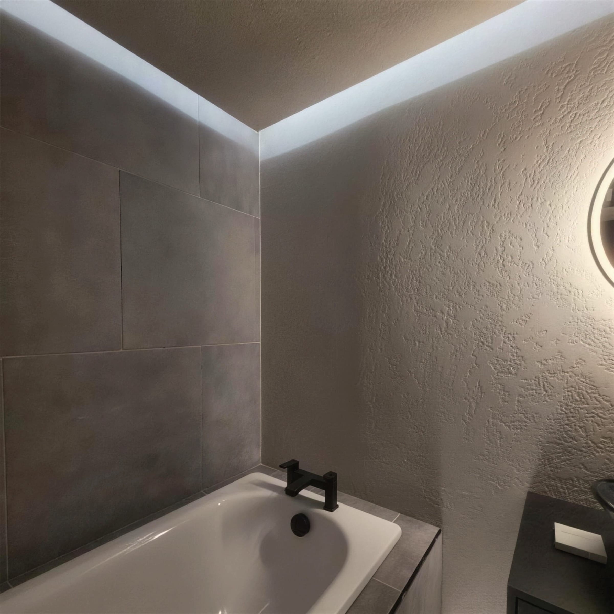 Finished bathroom corner: pitted travertine-effect plaster wall, perimeter LED cove, bath with matte black filler tap.