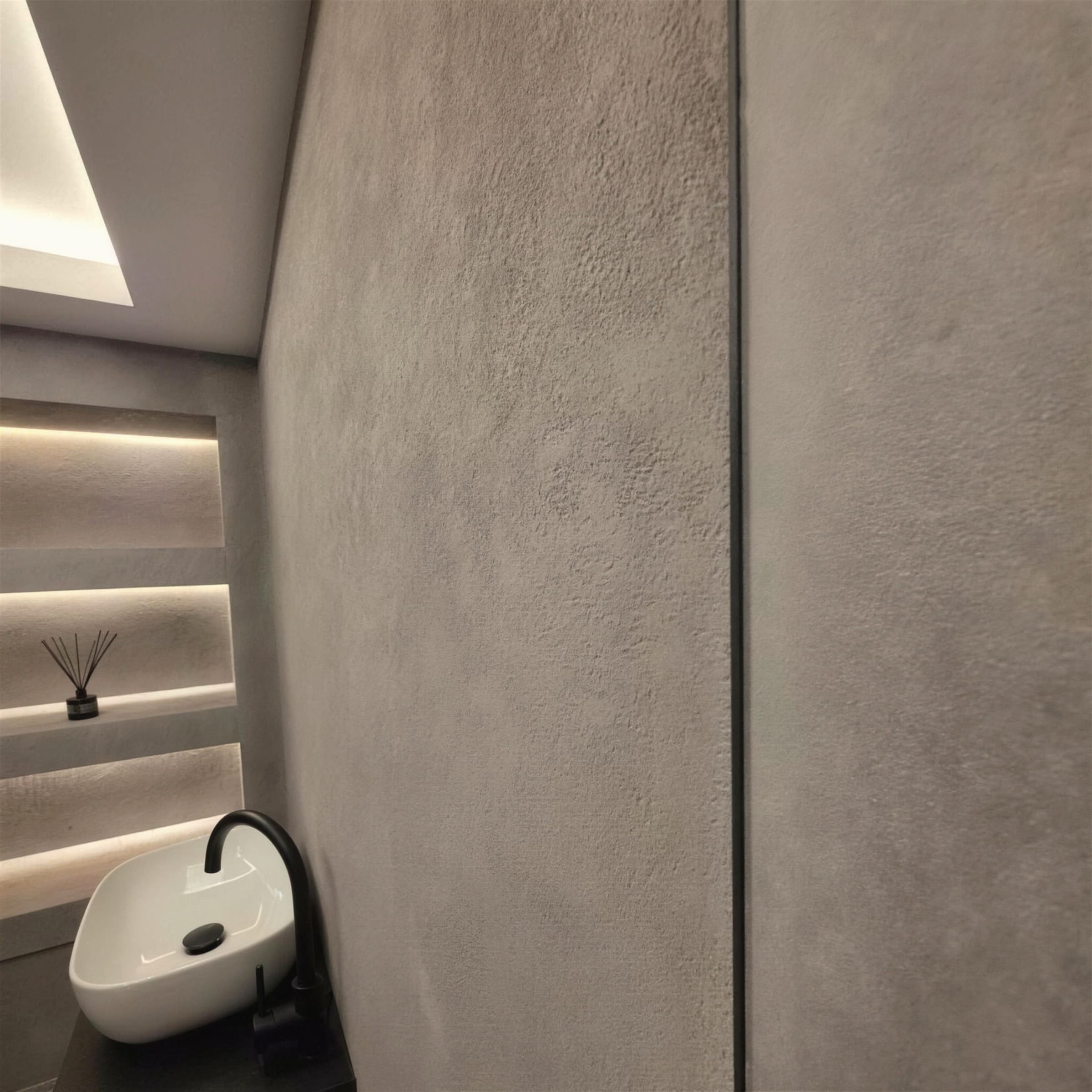 Close detail of a suede-textured grey plaster wall, warm LED niches and countertop basin softening the frame.