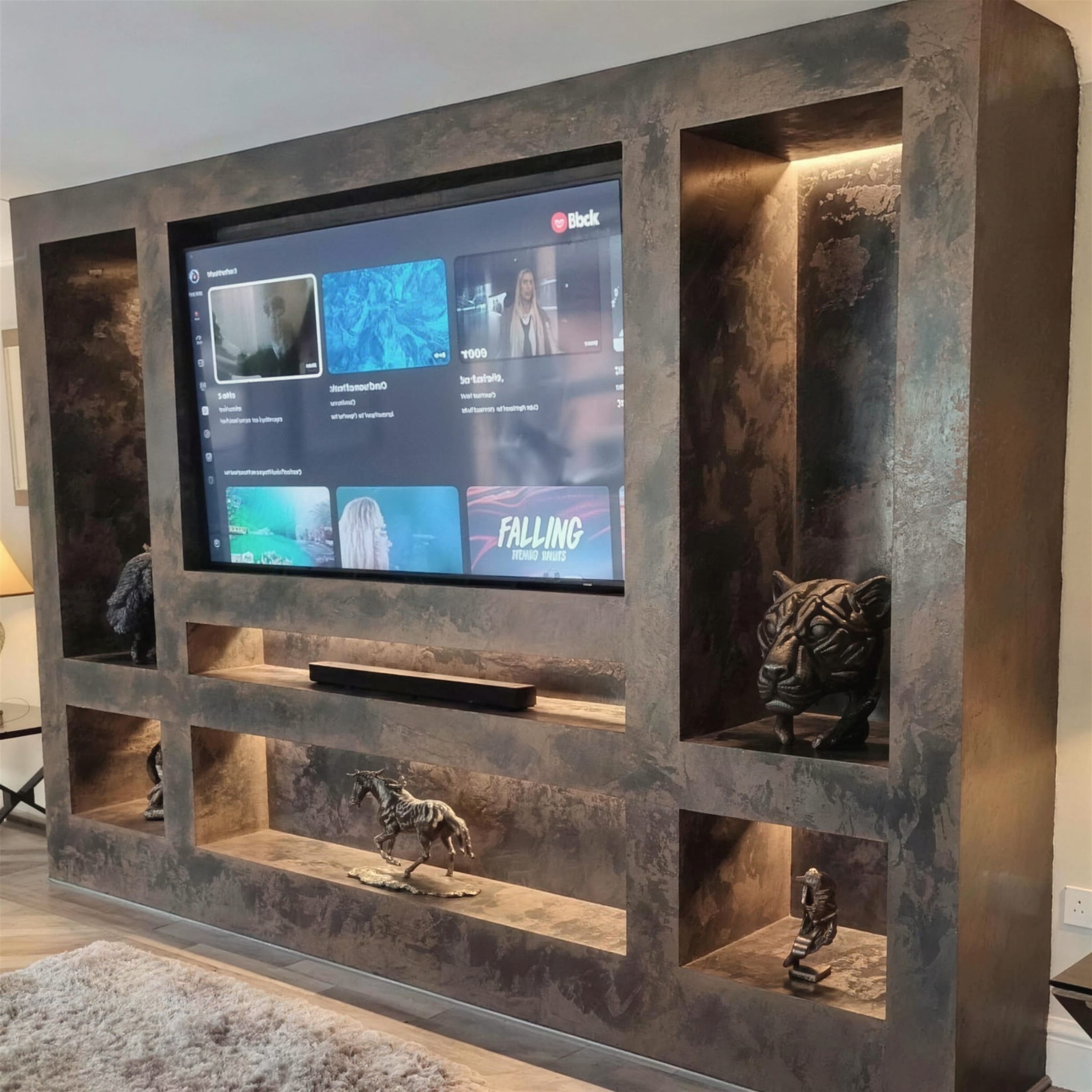 Sculptural media wall in mottled bronze metallic plaster, LED-lit niches with bronze figures; TV menu screen on.