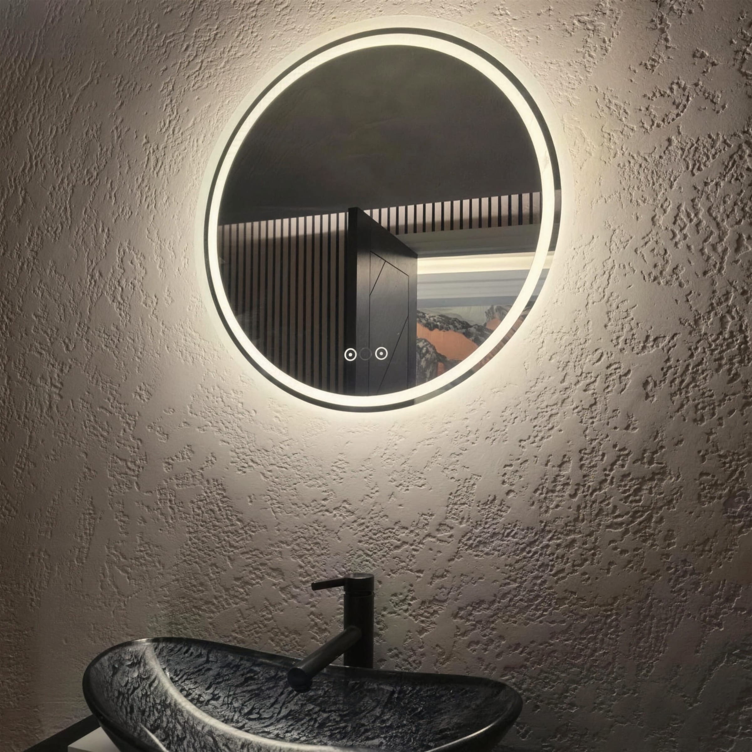 Warmly lit stippled plaster wall carrying a glowing round LED mirror, carved stone basin cropped at the base.