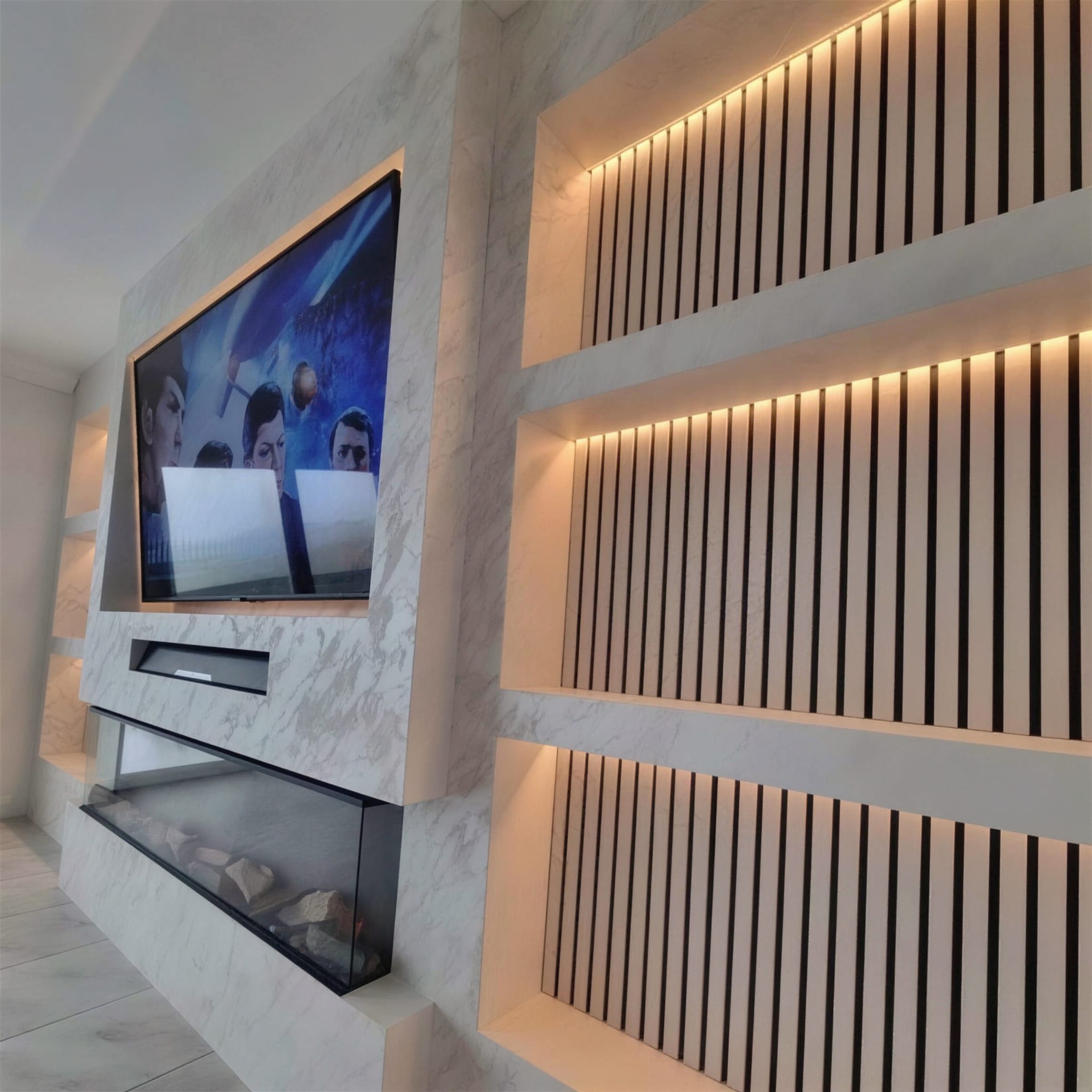 Bright media wall in white marble-effect plaster, warm LED shelving over black slats, inset fire below the TV.