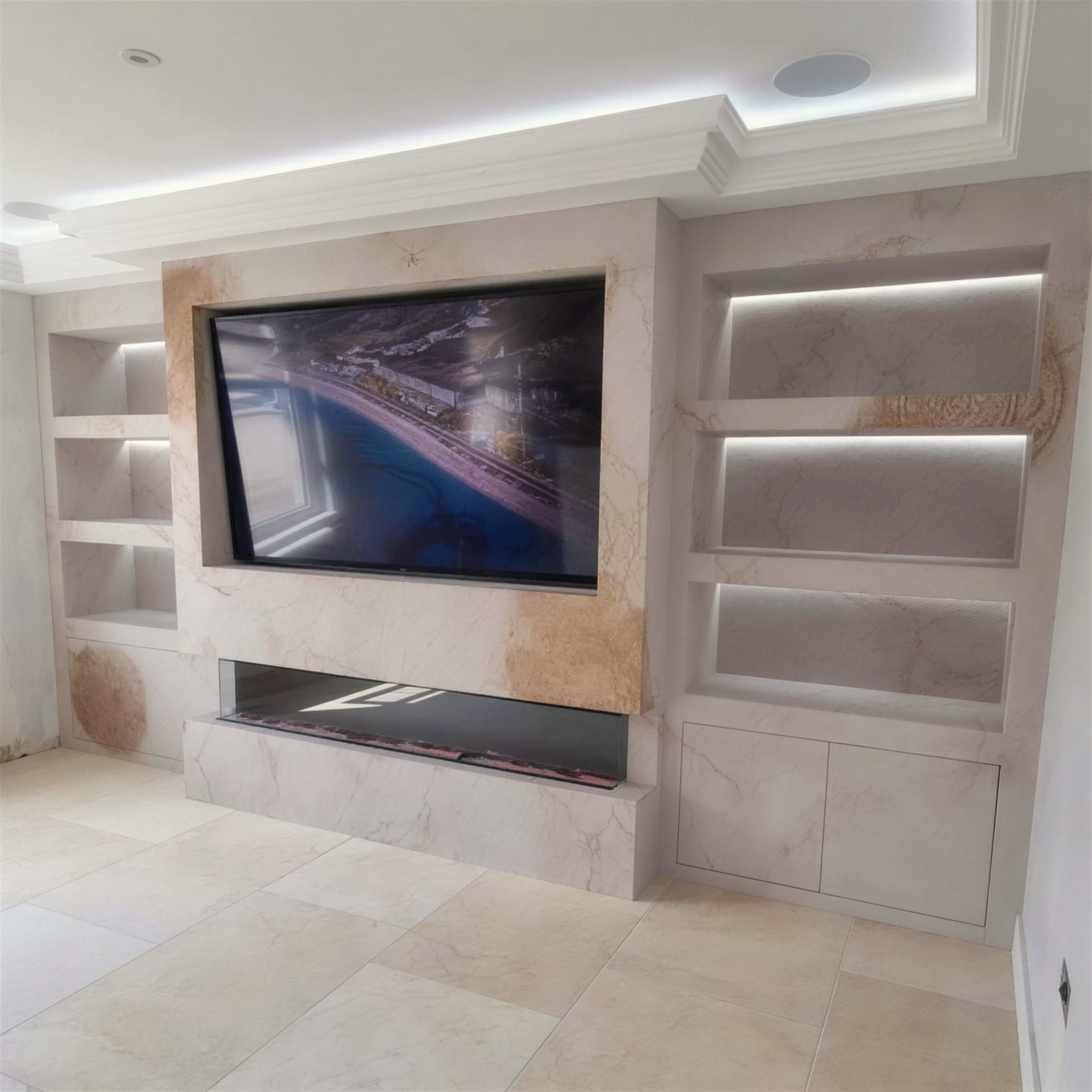 Media wall finished in cream travertine-effect plaster with rust-gold veining, LED-lit display niches, inset TV and linear fireplace over a travertine floor.