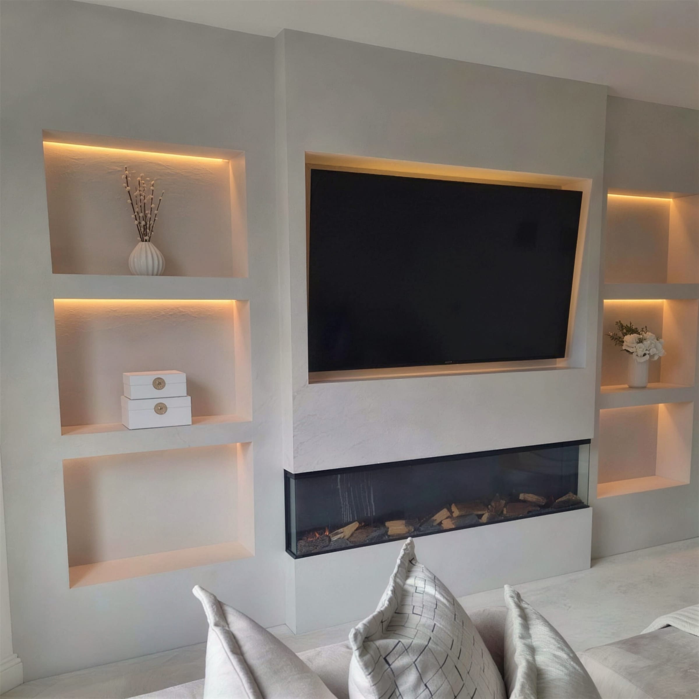 Styled media wall in smooth polished plaster with warm LED-lit niches holding vases and flowers, recessed TV and linear fireplace, cushions in the foreground.